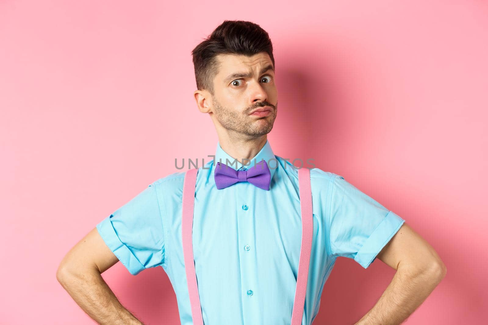 Image of young man with funny moustache and bow-tie standing in confident pose, staring with disbelief and doubts at camera, posing over pink background.