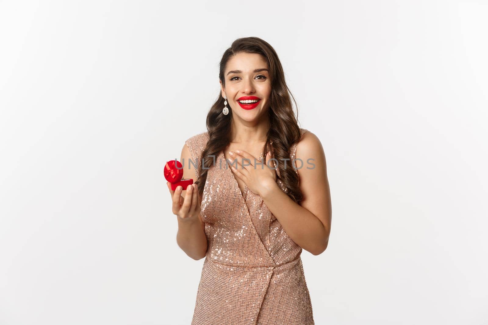 Image of beautiful woman in glamour dress receiving engagement ring, looking surprised and happy at camera, being proposed, standing over white background.