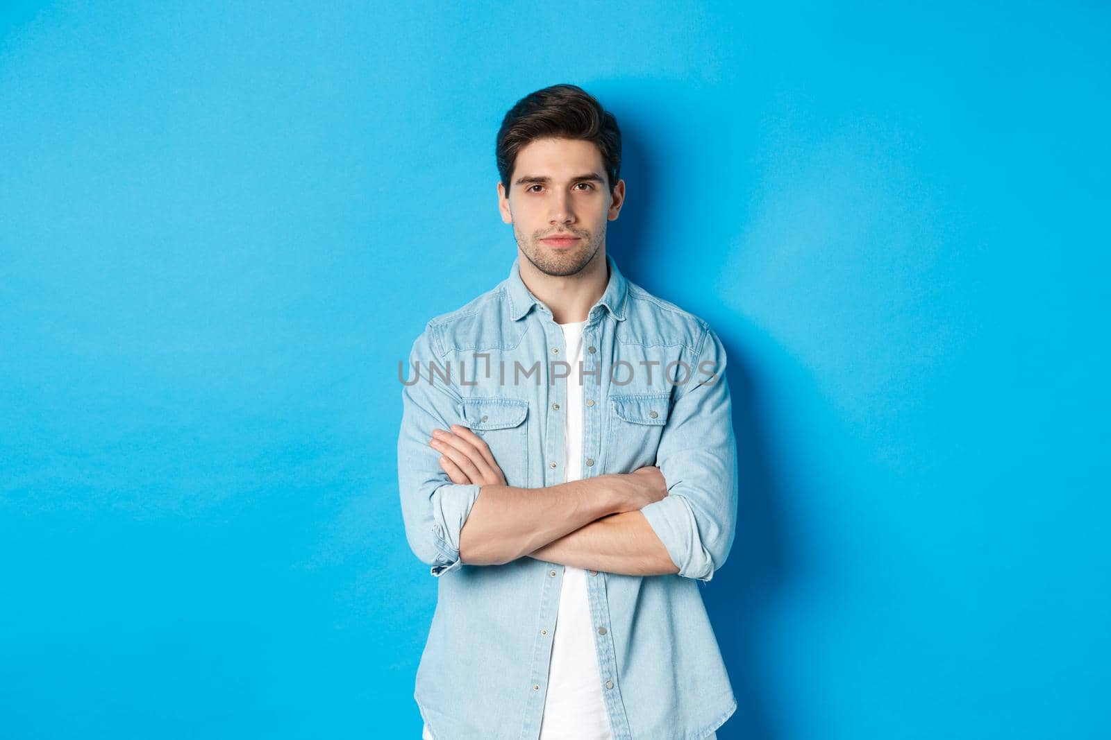 Image of handsome caucasian man in casual outfit, looking serious and confident, standing against blue background.