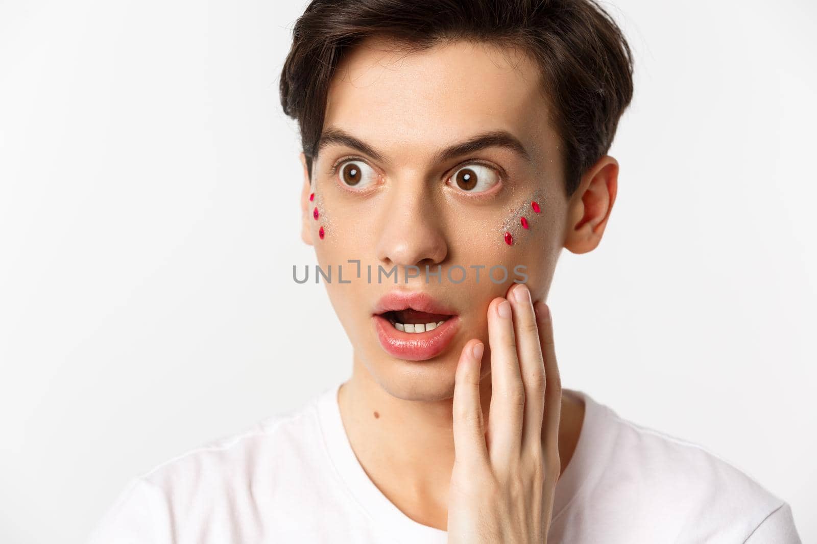 People, lgbtq and beauty concept. Headshot of gay man with glitter on face, looking left in awe, express surprise and disbelief, standing against white background.