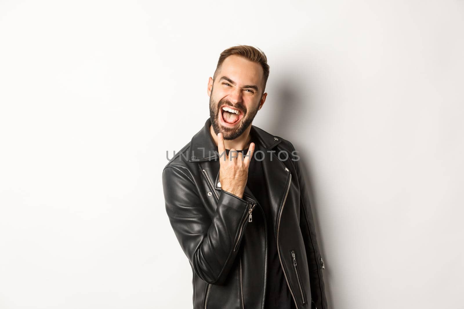 Cool adult man in black leather jacket, showing rock on gesture and tongue, enjoying music festival, standing over white background.