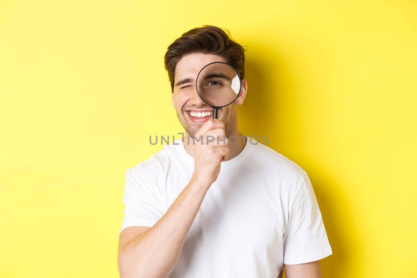 Close-up of young man looking through magnifying glass and smiling, searching something, standing over yellow background by Benzoix
