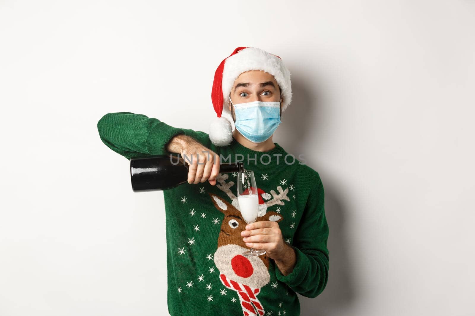 Concept of covid-19 and Christmas holidays. Happy guy in face mask and santa hat, celebrating New Year party and pouring glass of champagne, white background.