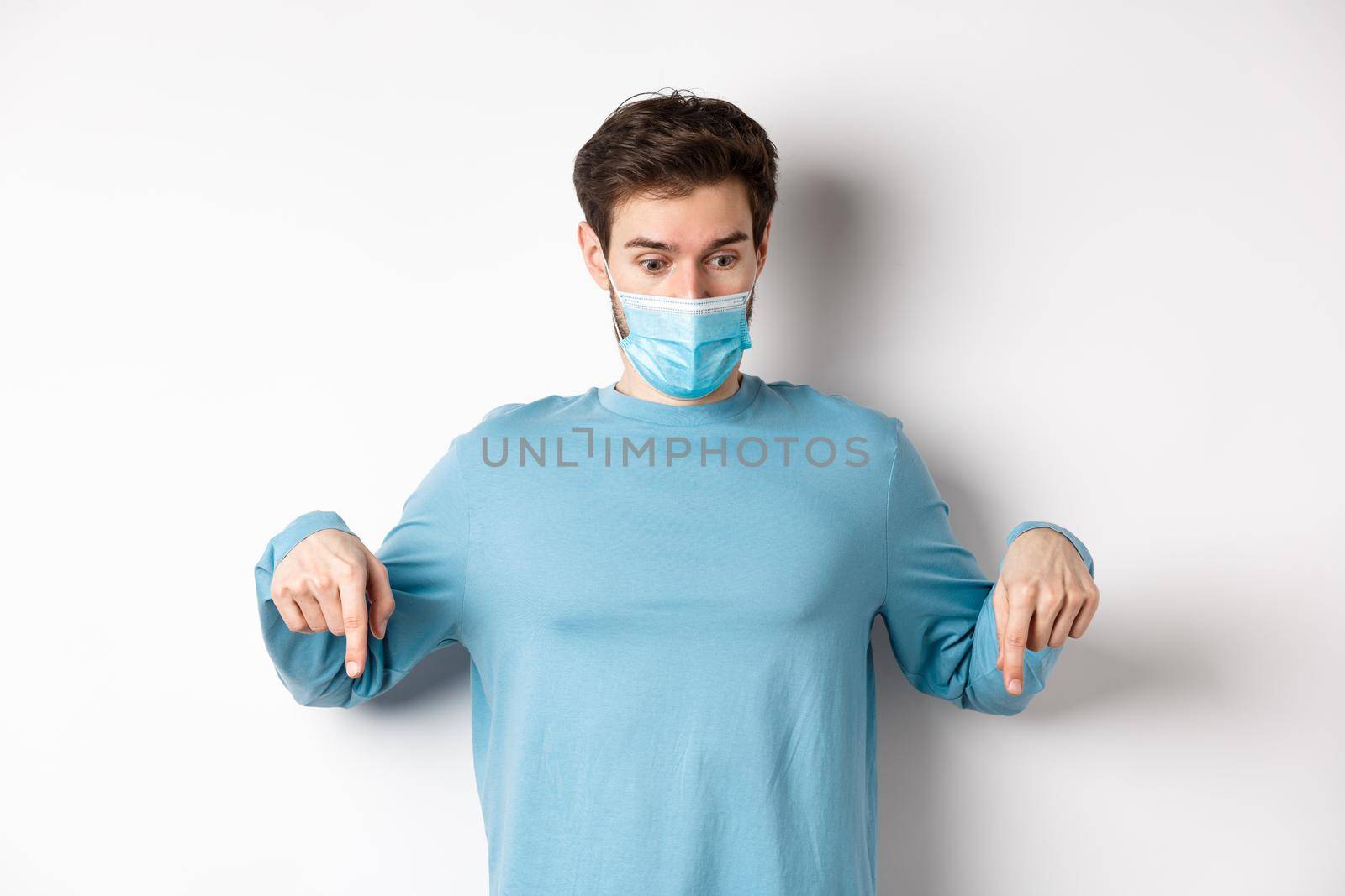 Covid-19, health and quarantine concept. Curious man checking out advertisement, pointing and looking down, wearing medical mask with casual blue sweatshirt, white background.