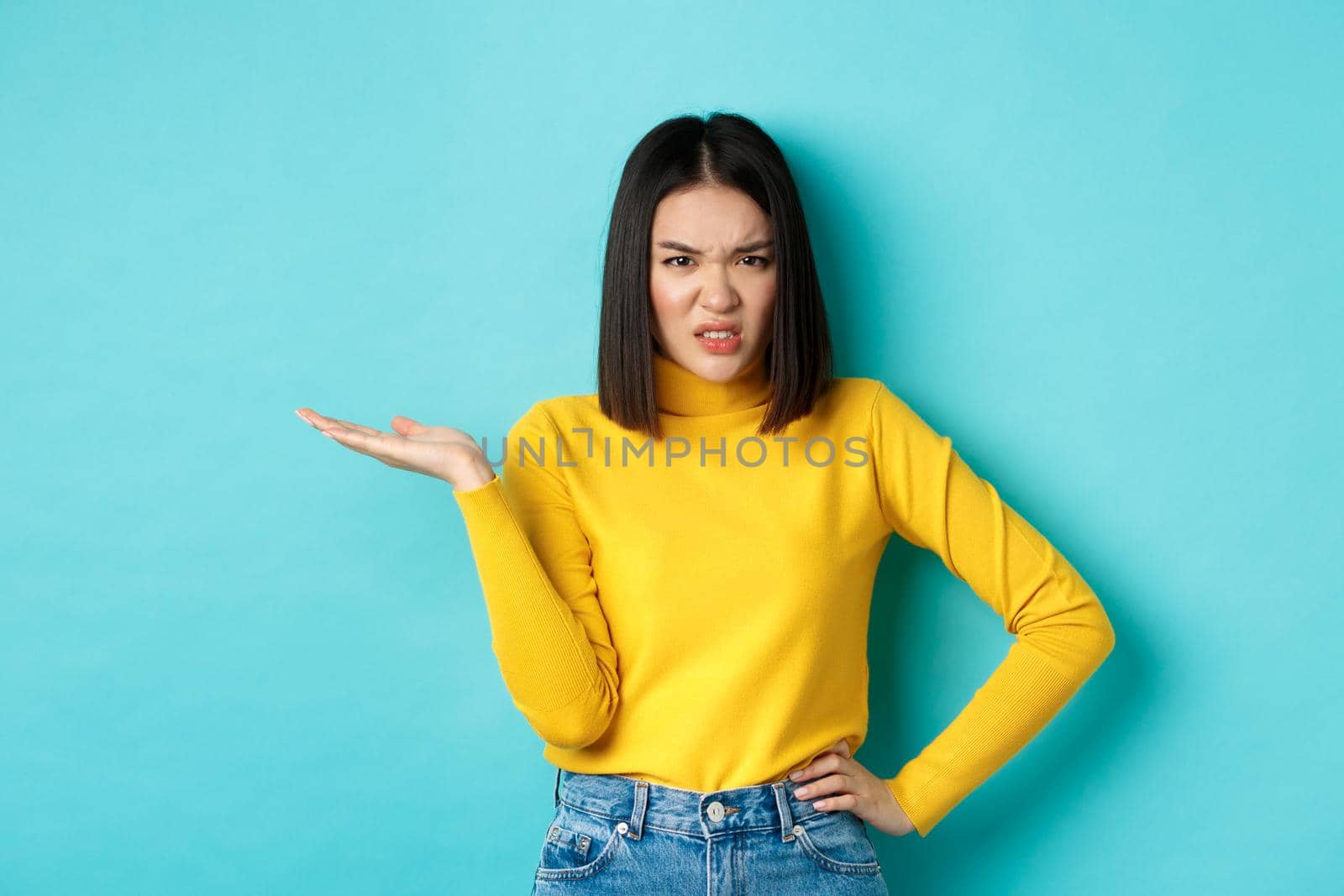Frustrated and pissed-off asian girl raising hand up and grimacing bothered, staring at something with annoyance and disappointment, standing over blue background.