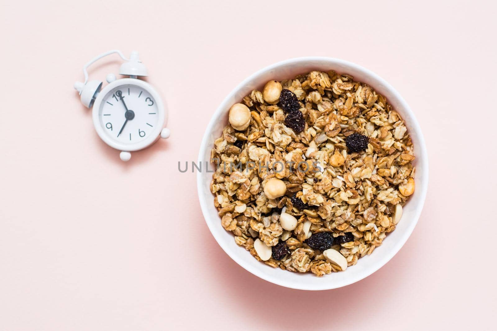 Healthy eating. Baked granola of oats, nuts and raisins in a bowl and an alarm clock on a pink background. Top view by Aleruana