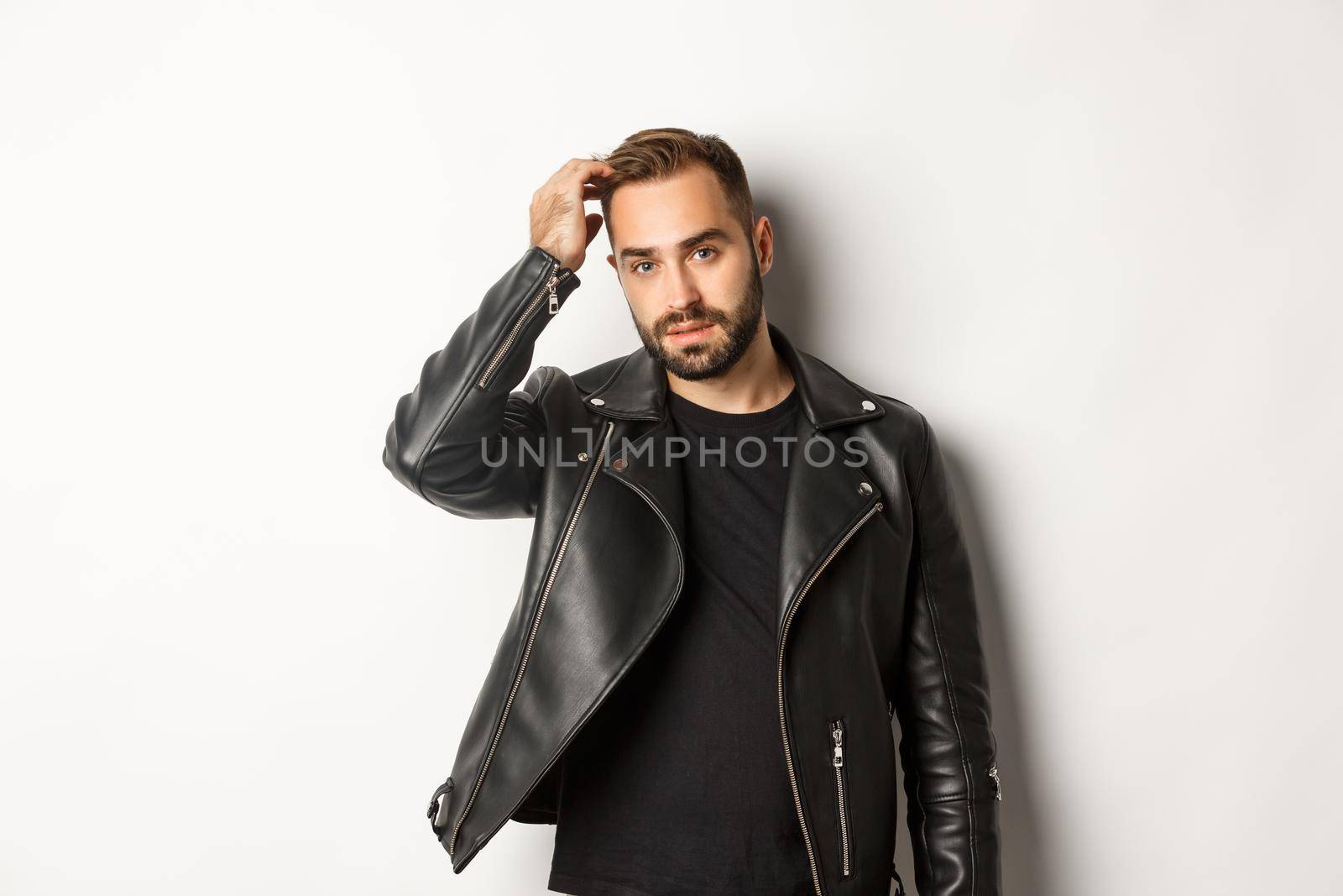 Handsome macho man in black biker jacket, touching his haircut and looking cool, white background.