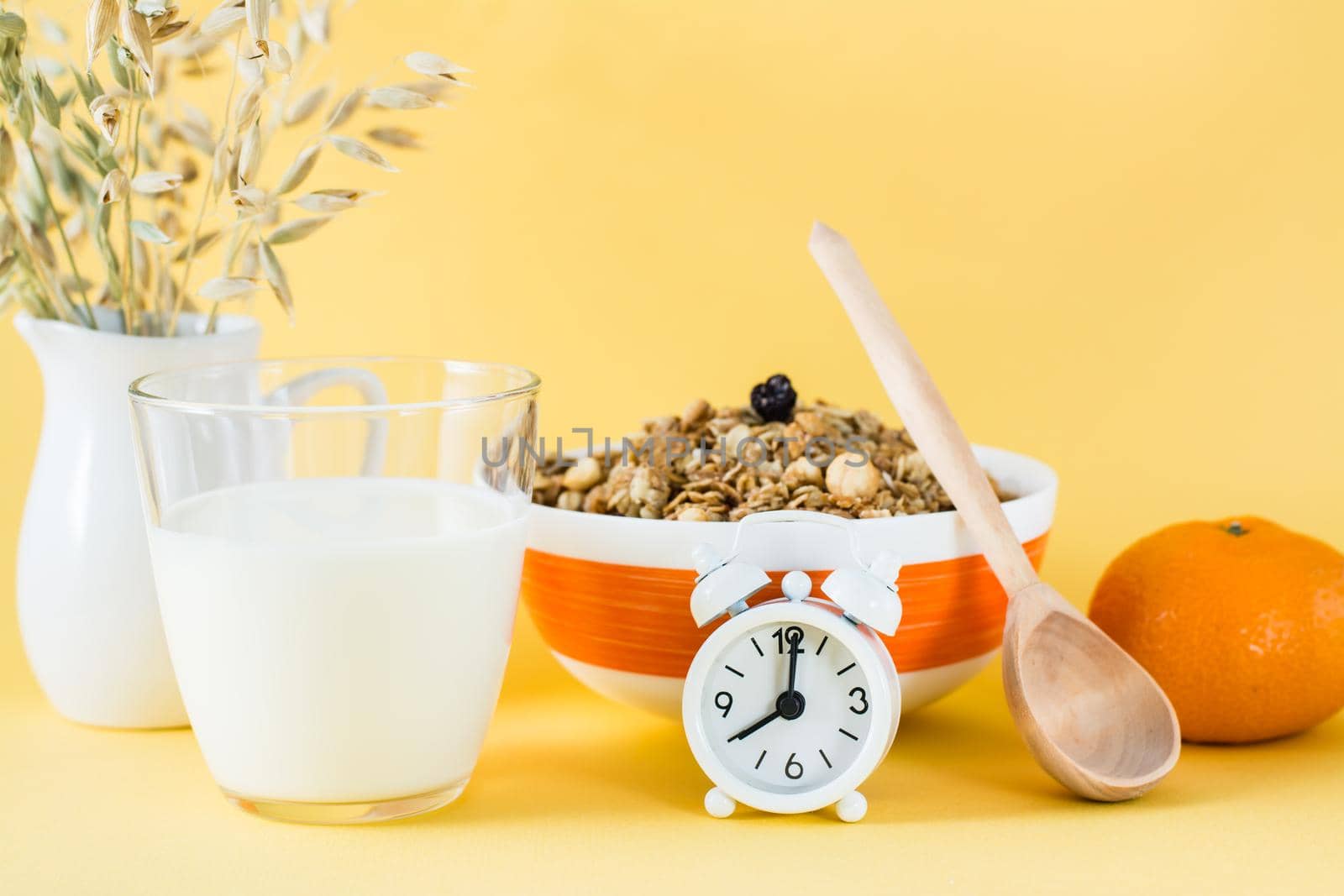 Healthy hearty breakfast. Baked granola in bowl, glass of milk, orange and alarm clock on yellow background