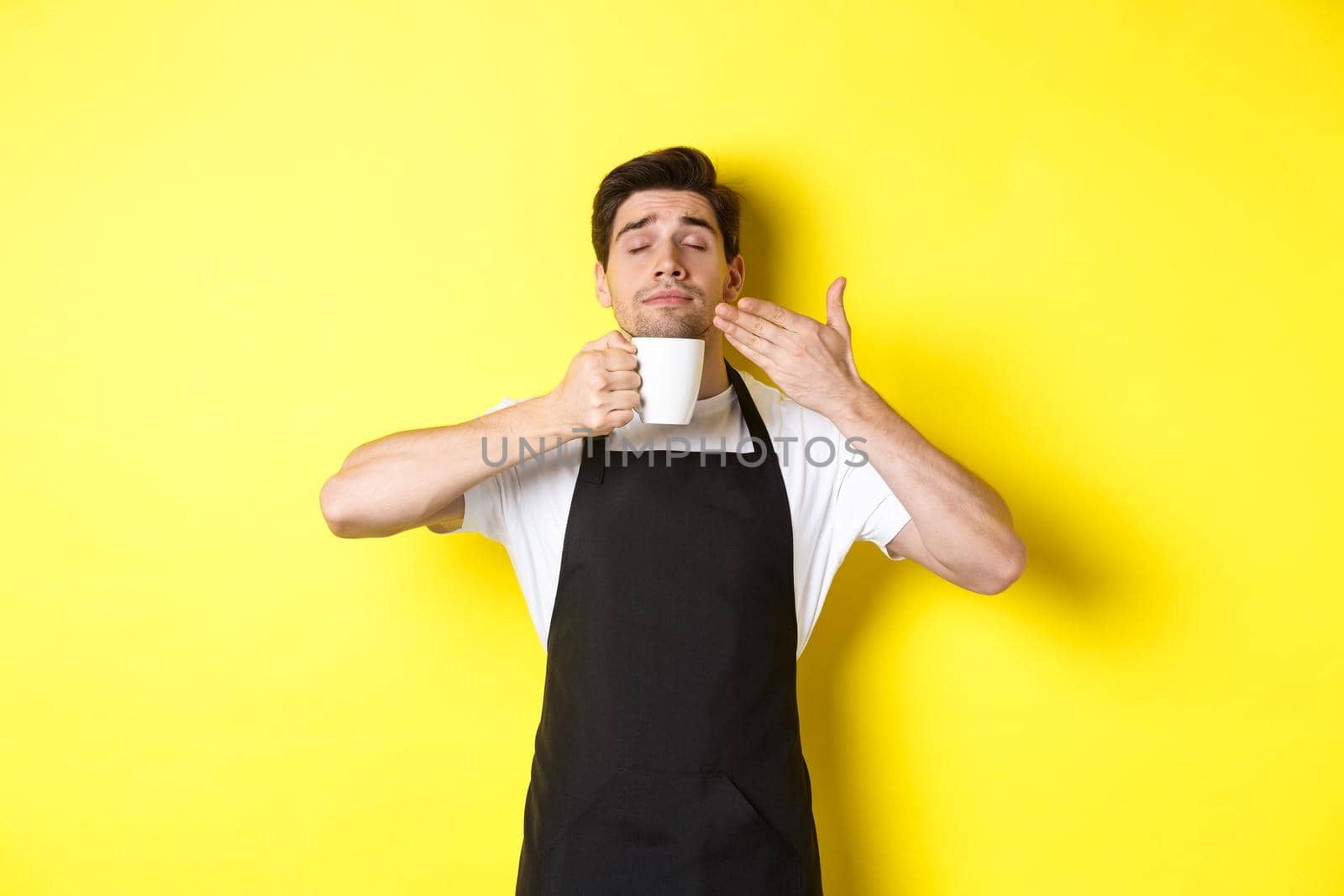 Barista enjoying smell of coffee in mug, standing pleased with eyes closed, wearing black apron by Benzoix