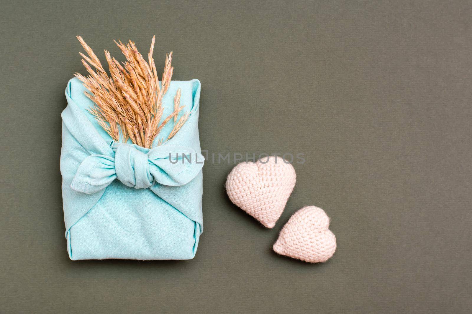 Eco friendly furoshiki gift with ears of dry grass and two knitted hearts on a green background by Aleruana