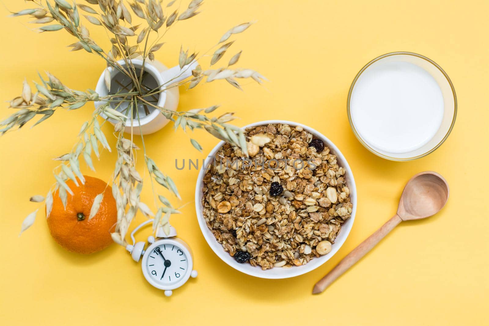 Healthy hearty breakfast. Baked granola in bowl, glass of milk, orange, wooden spoon and alarm clock on yellow background. Top view