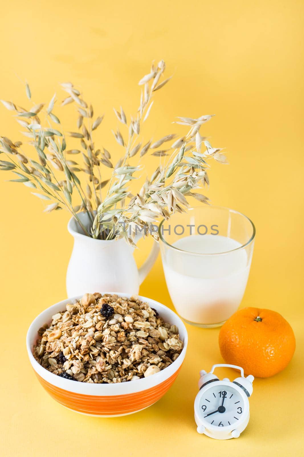 Healthy hearty breakfast. Baked granola in bowl, glass of milk, orange and alarm clock on yellow background. Vertical view
