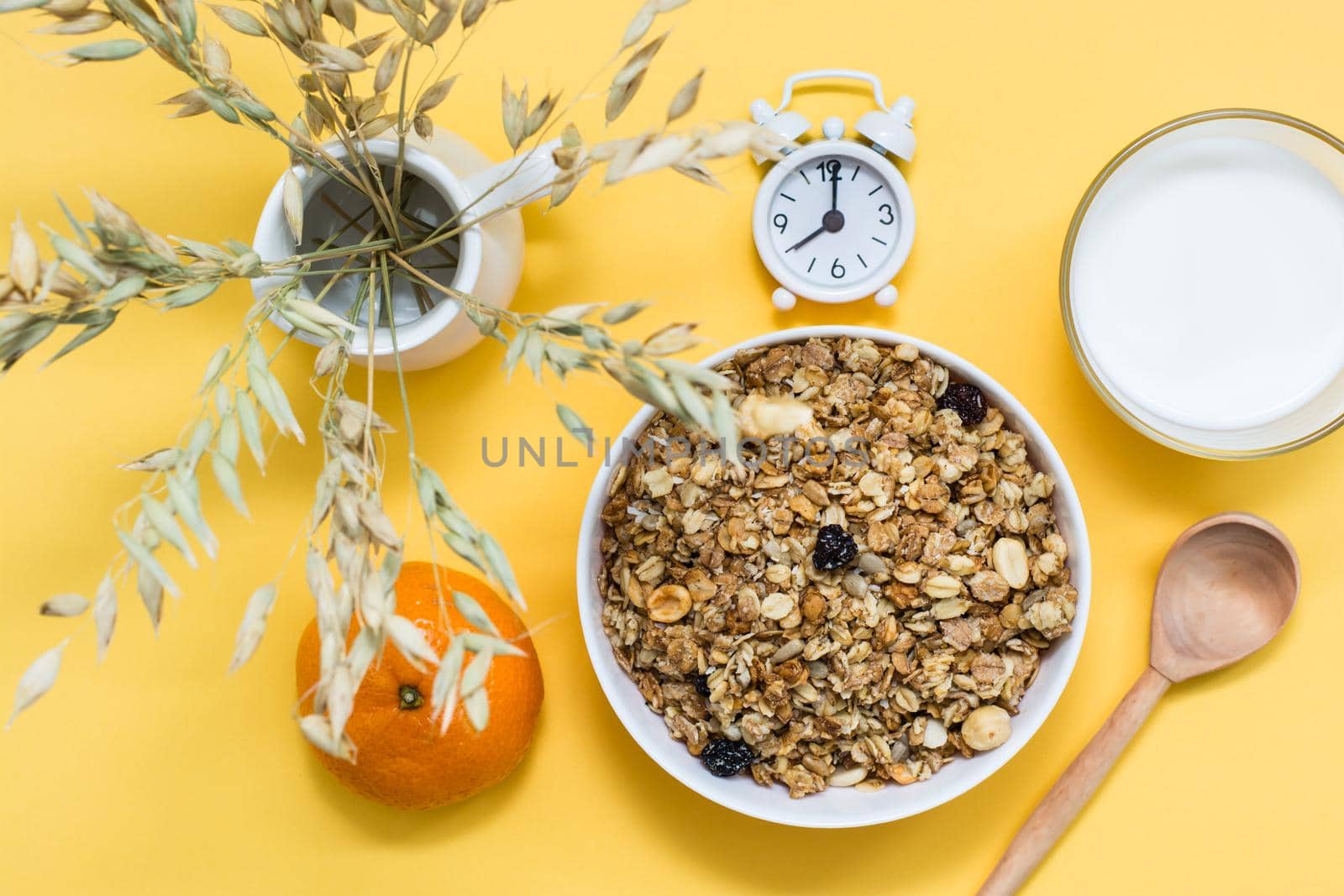 Healthy hearty breakfast. Baked granola of oats, nuts and raisins in a bowl, a glass of milk, an orange and an alarm clock on a yellow background. Top view by Aleruana