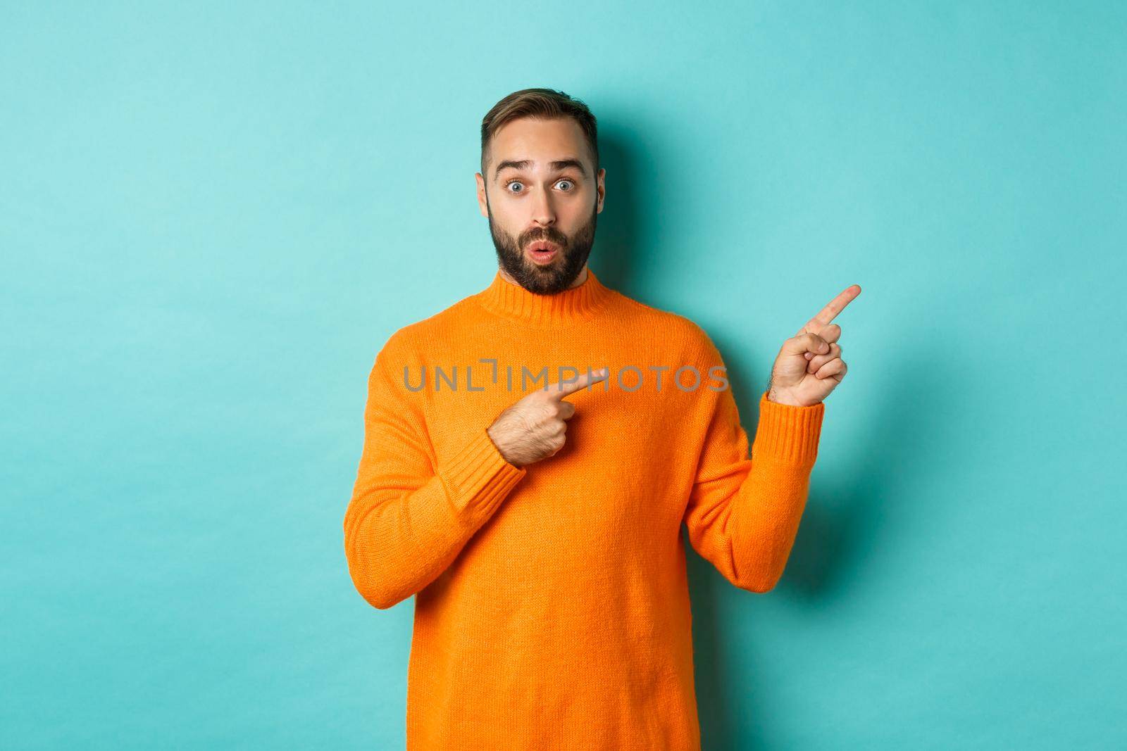 Amazed guy showing advertisement, pointing fingers right at banner, standing against turquoise background.