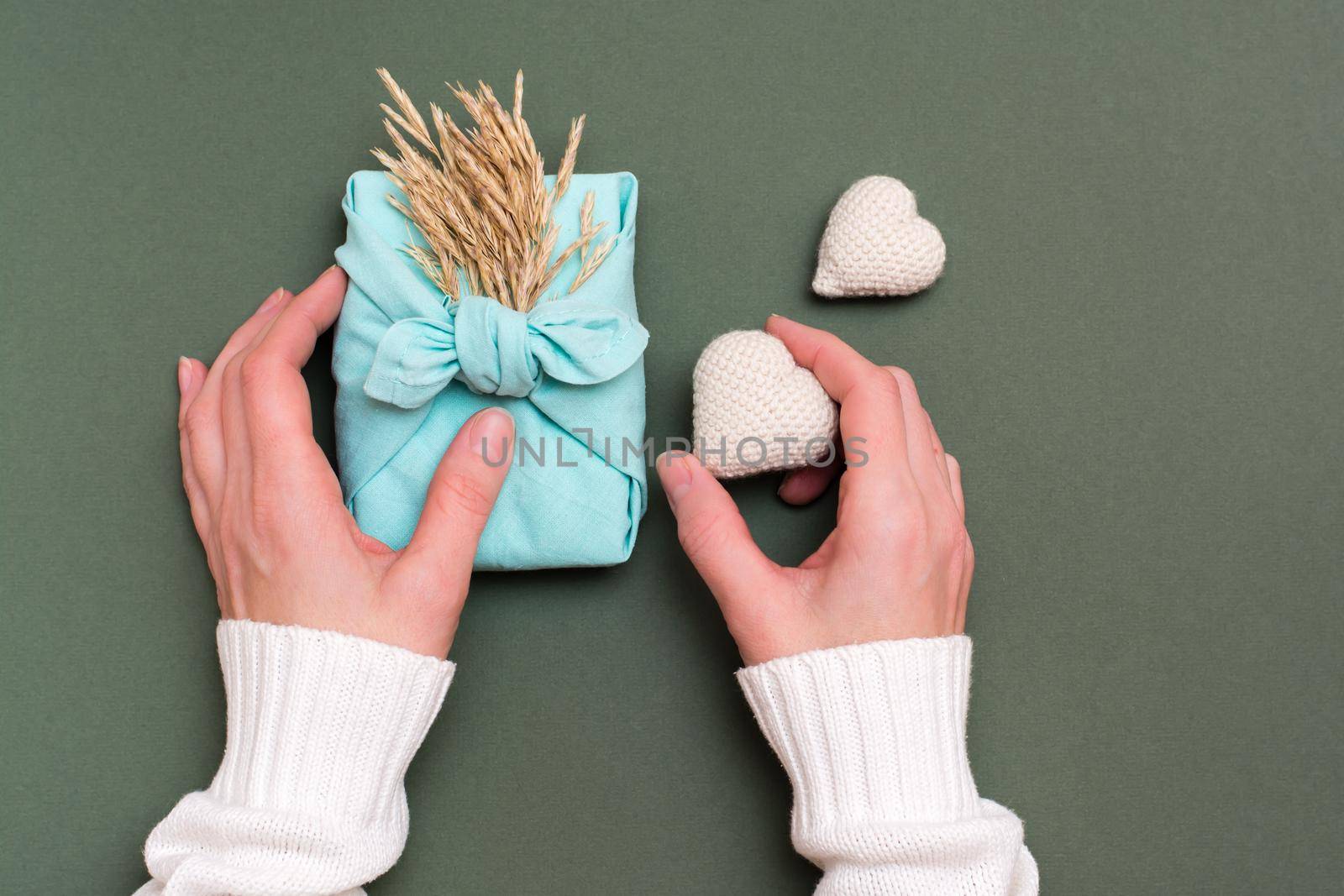 Female hands hold eco friendly furoshiki gift with ears of dry grass and two knitted hearts on a green background