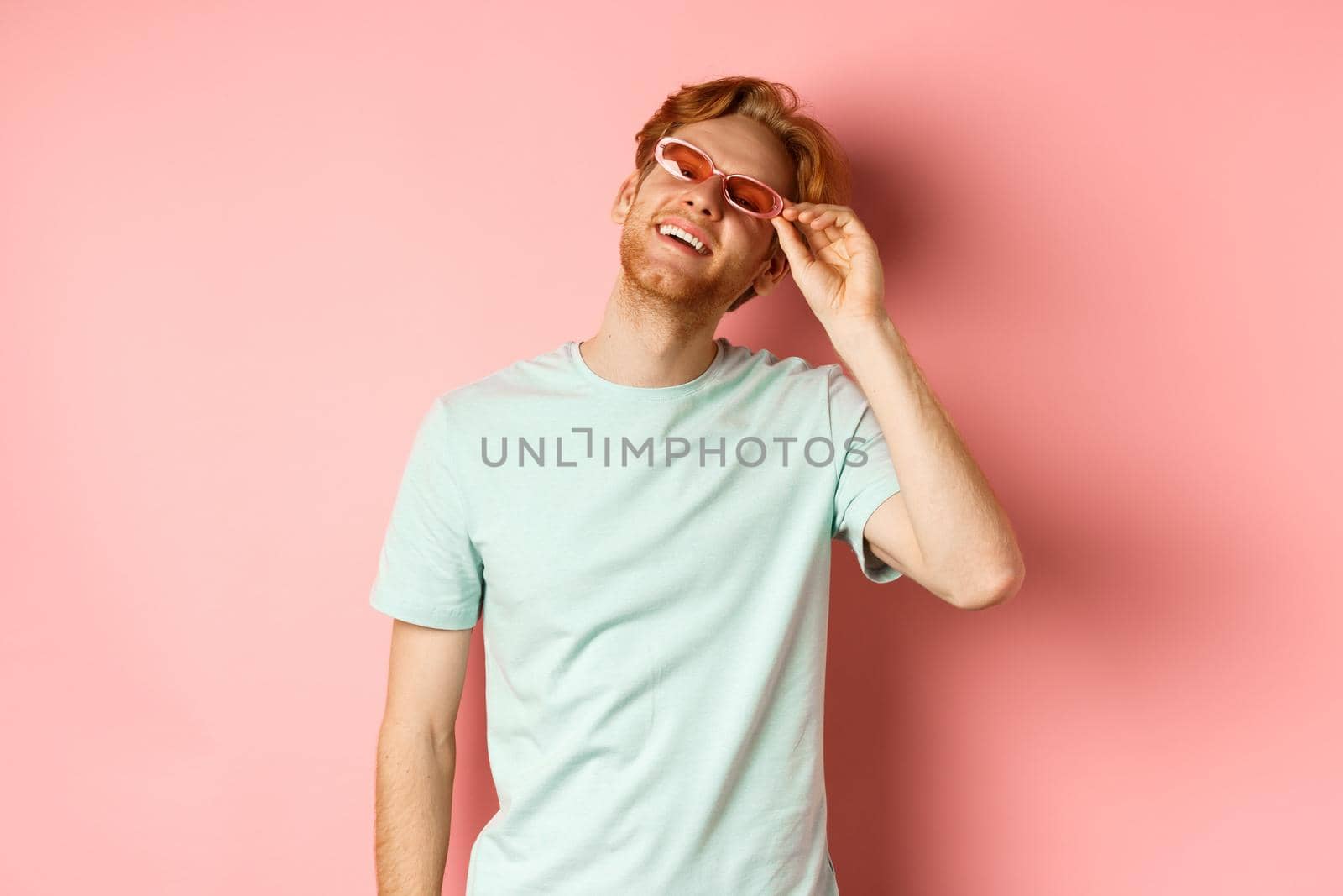 Tourism and vacation concept. Happy young man with red hair relaxing, wearing sunglasses and smiling carefree, standing over pink background by Benzoix