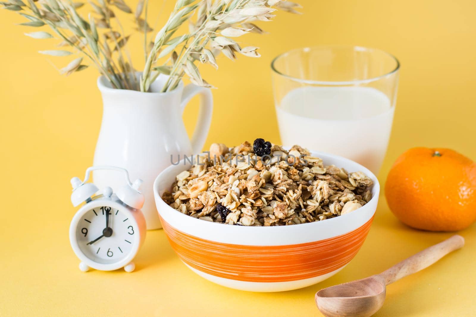 Healthy hearty breakfast. Baked granola of oats, nuts and raisins in a bowl, a glass of milk, an orange and an alarm clock on a yellow background