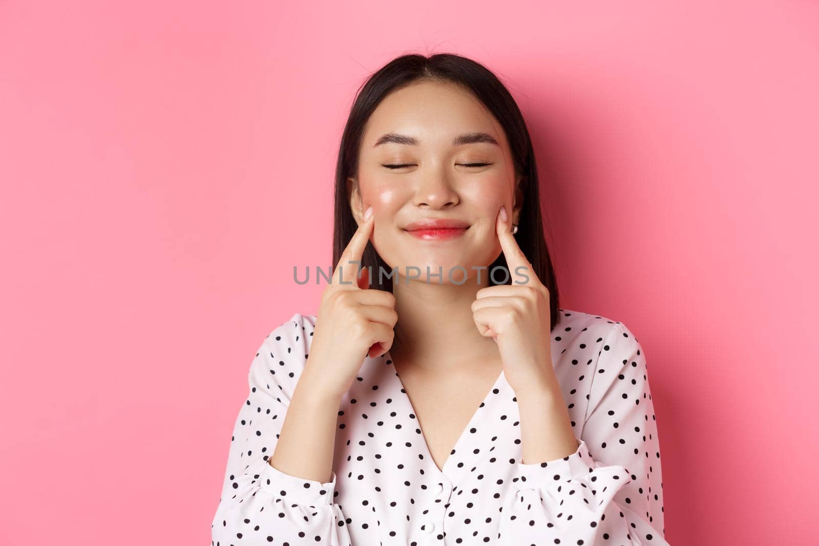 Beauty and lifestyle concept. Close-up of beautiful asian woman poking cheeks with closed eyes, smiling satisfied, standing over pink background by Benzoix