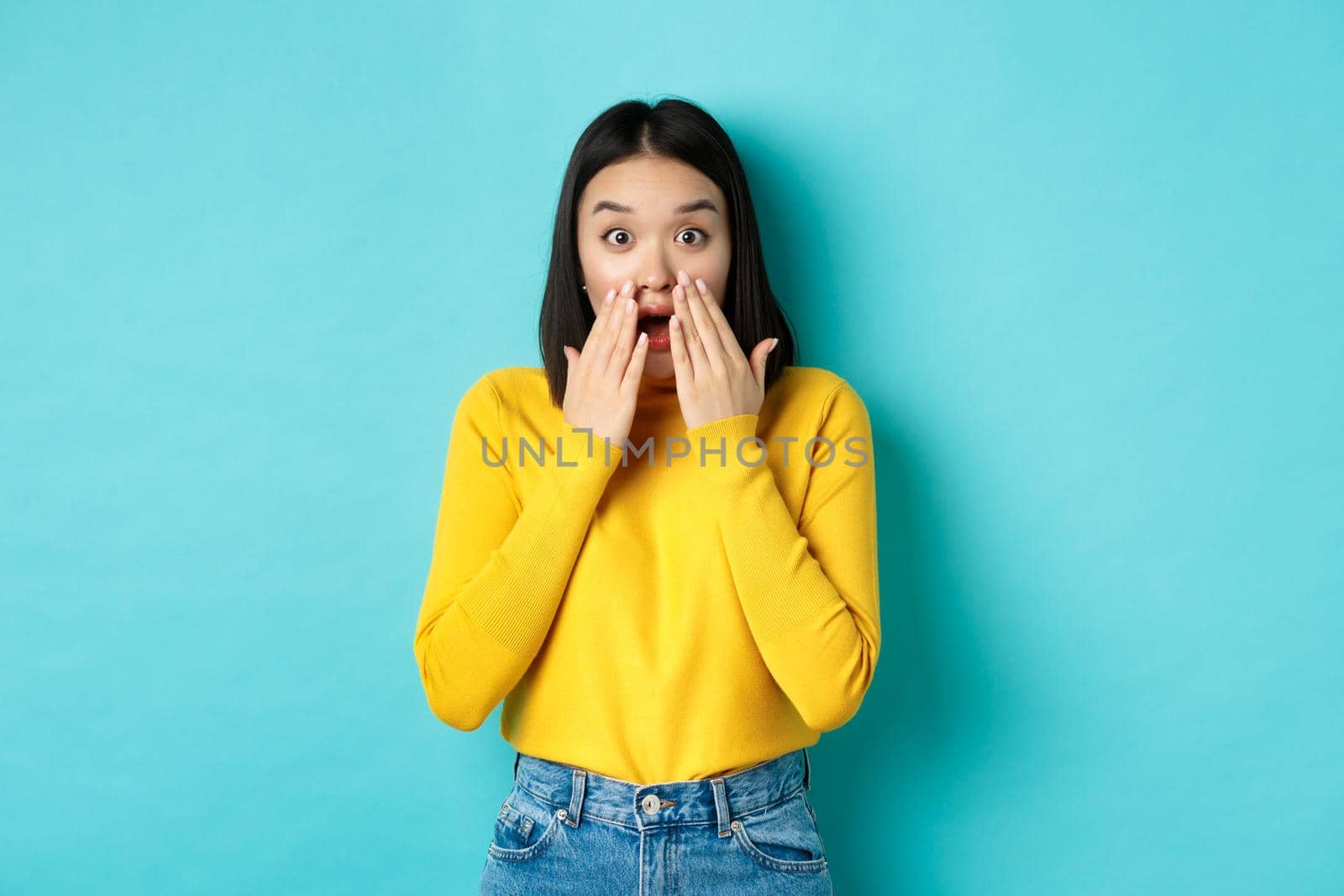 Portrait of impressed korean girl saying wow, gasping and staring amazed at camera, standing in yellow pullover over blue background.