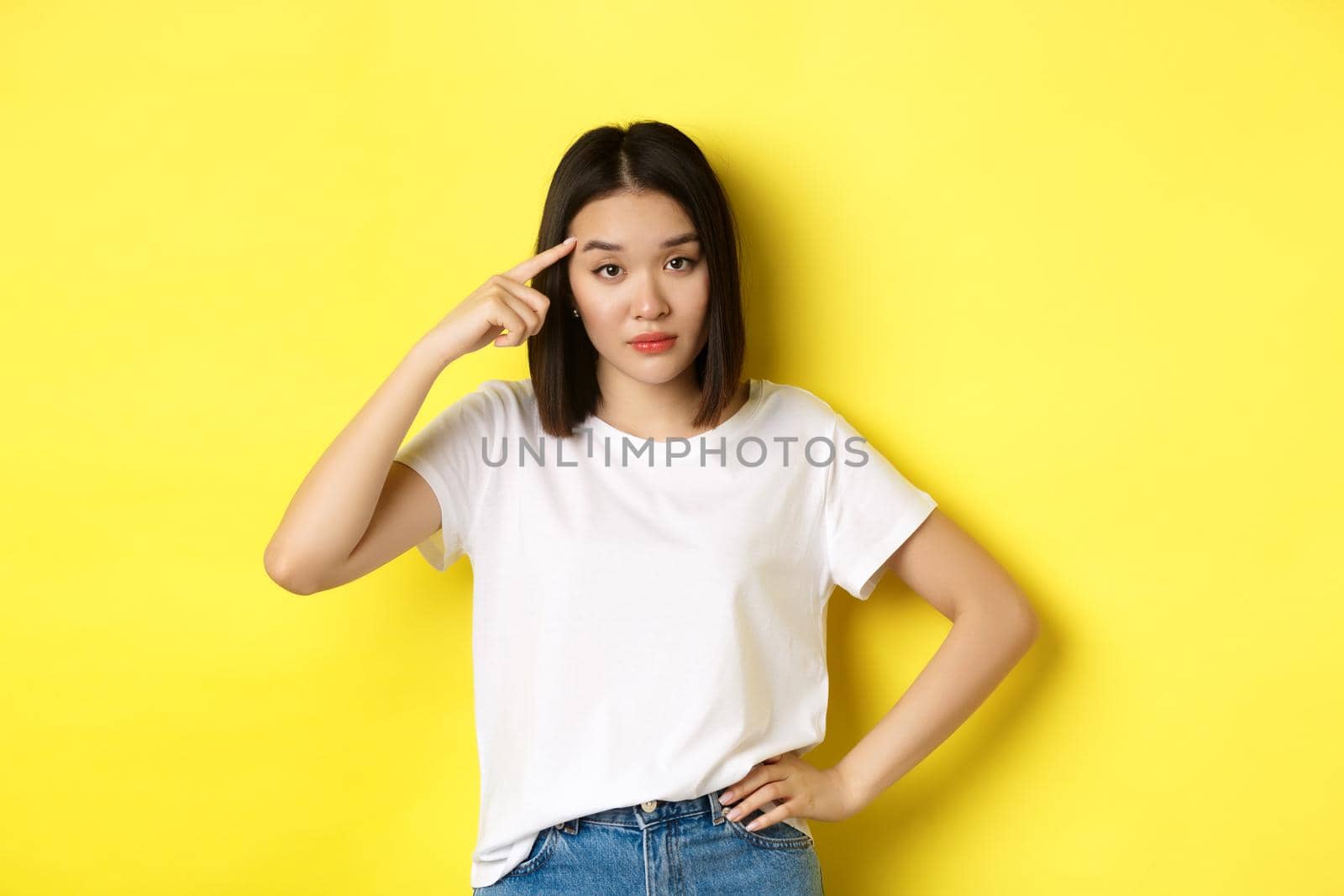 Annoyed young asian woman scolding someone stupid, pointing finger at head, asking are you crazy, standing over yellow background.