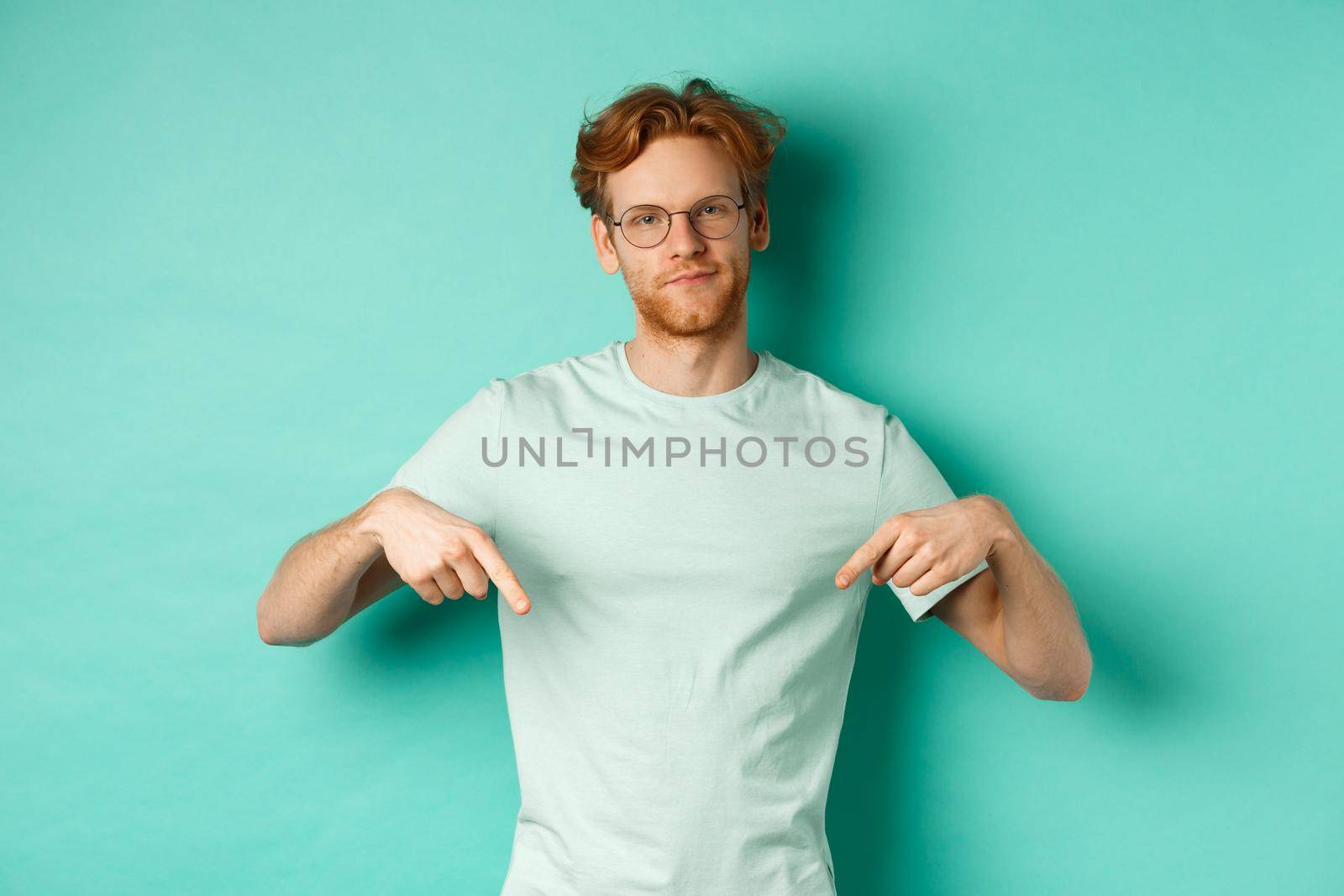 Skeptical and bothered man with ginger hair and beard, wearing glasses and t-shirt, smirk and point fingers down, showing promo with judgy face, turquoise background.