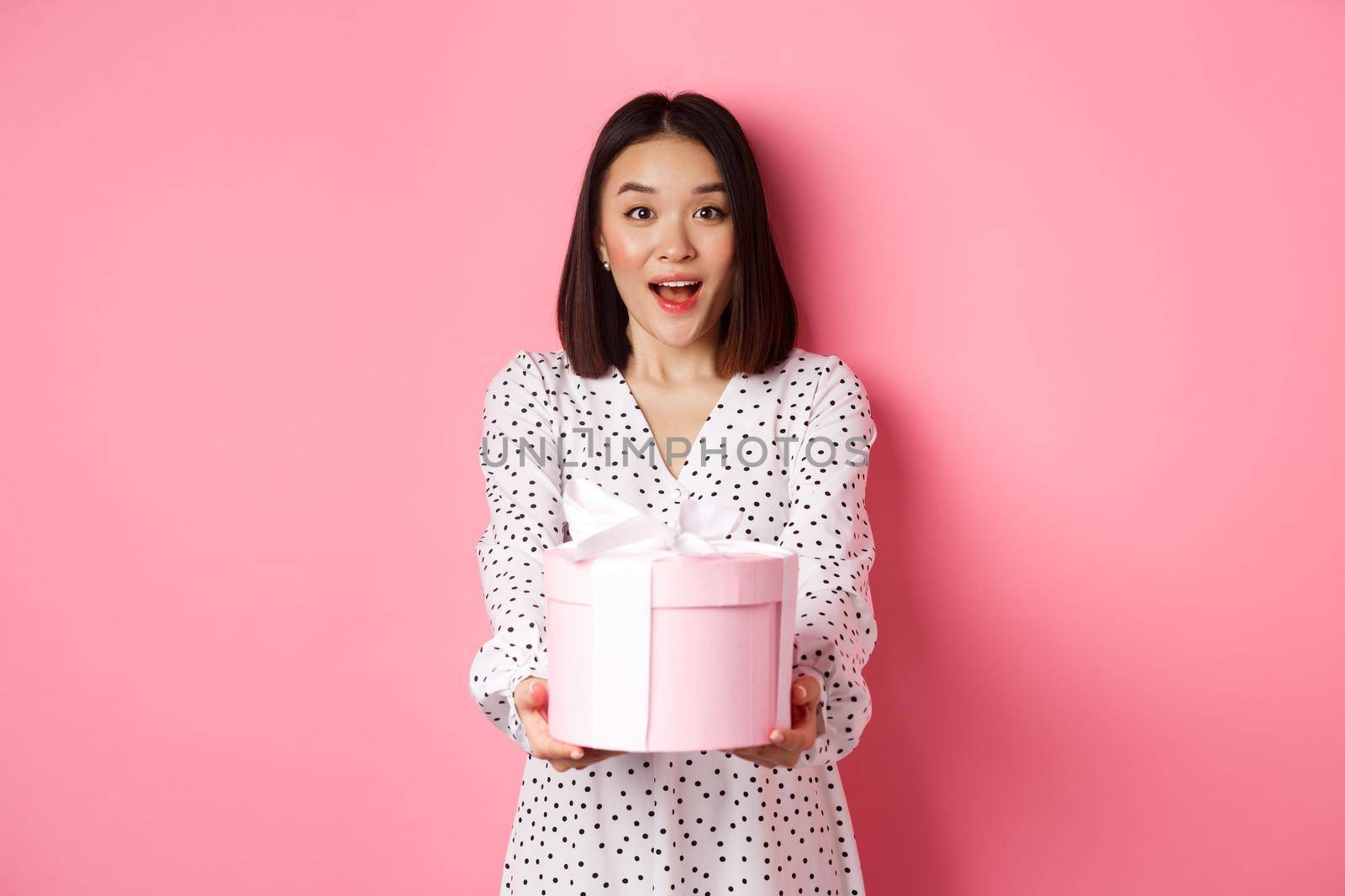 Cute asian woman congratulate with holiday or birthday, giving gift in cute box, standing over pink background.