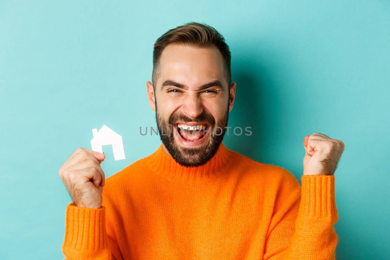 Real estate. Cheerful man buying apartment, rejoicing and saying yes, showing small paper house, standing over light blue background.