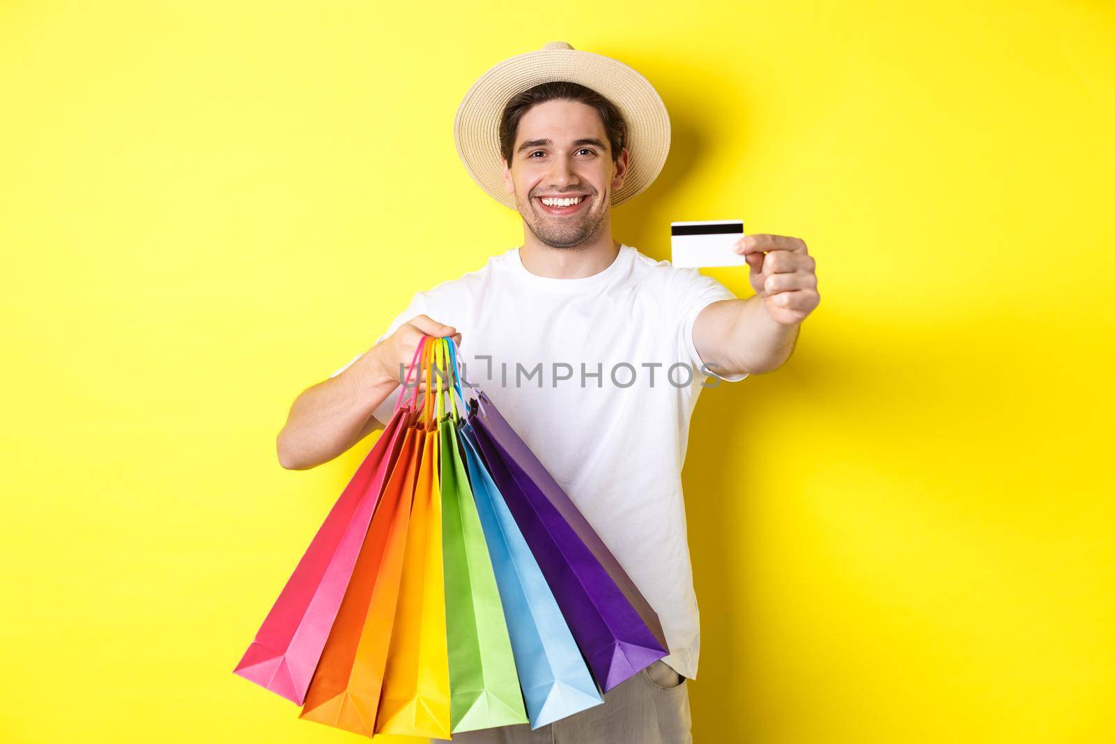 Smiling young man buying things with credit card, holding shopping bags and looking happy, standing over yellow background.