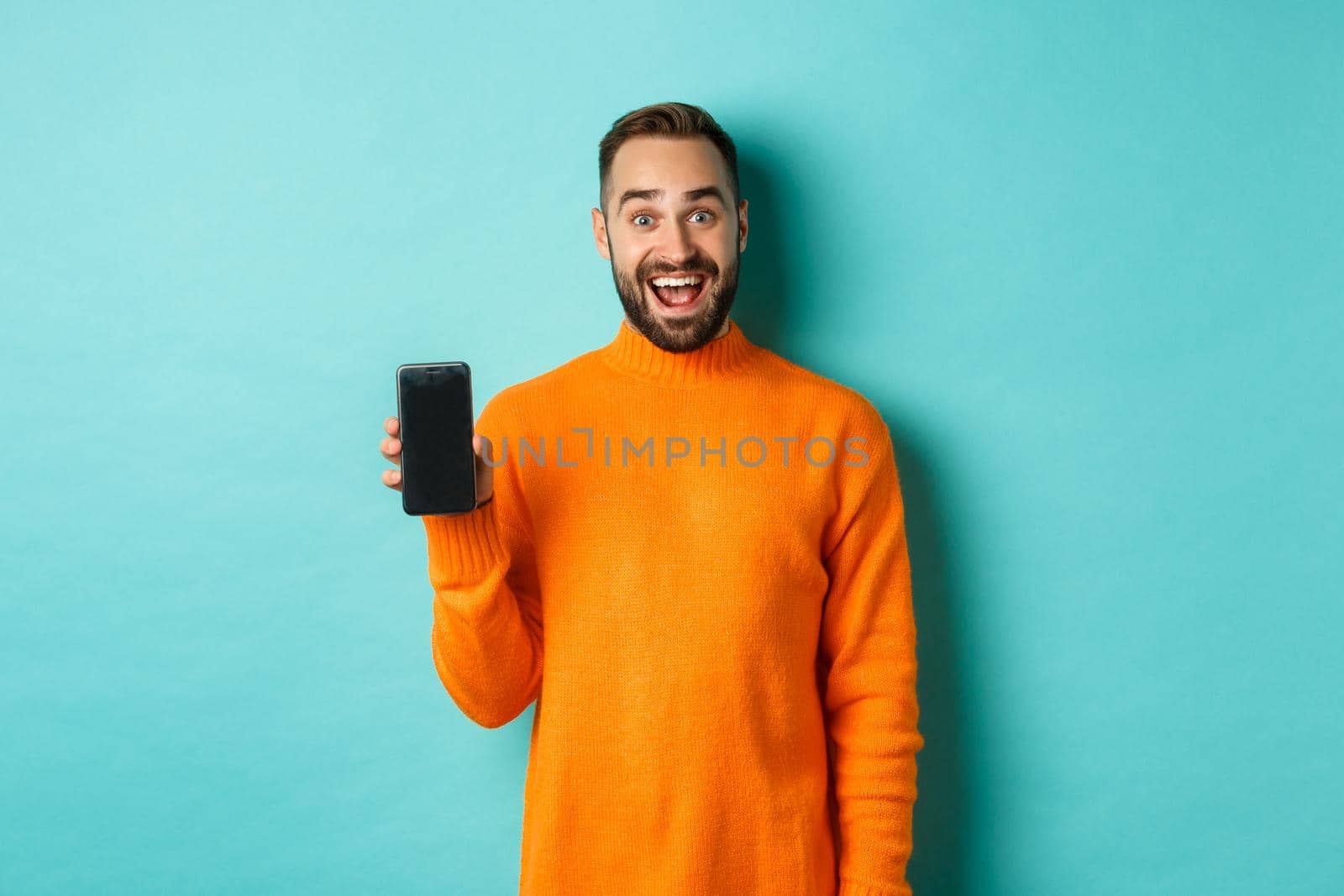 Photo of happy man showing mobile screen, introduce online store, application, standing over turquoise background by Benzoix