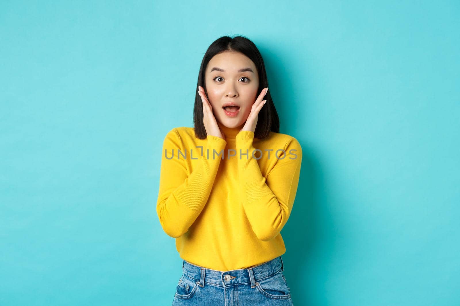 Portrait of surprised asian girl checking out promo, gasping amazed and touching cheeks, staring at camera amazed, blue background.