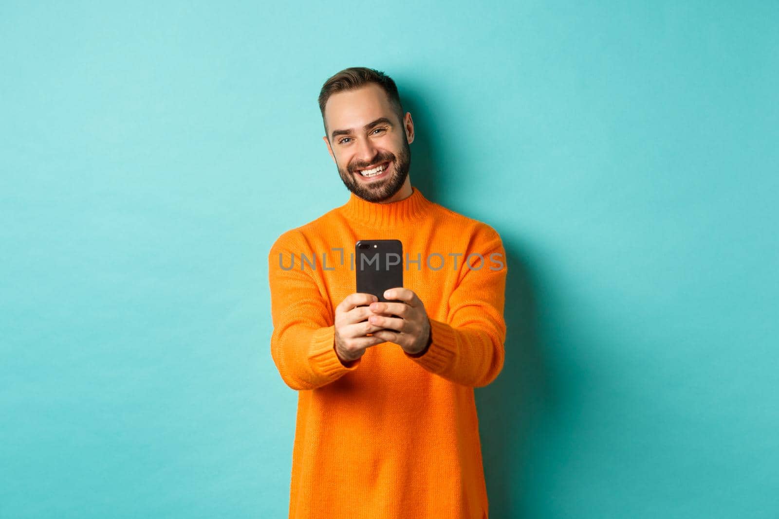 Happy handsome man taking photo on mobile phone, take pictures with smartphone camera, standing over light blue background.