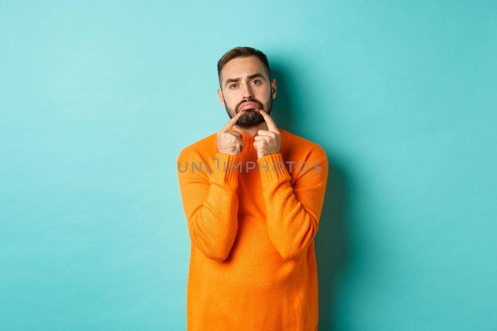 Image of gloomy bearded man, making sad face and frowning, standing upset in orange sweater against turquoise background.