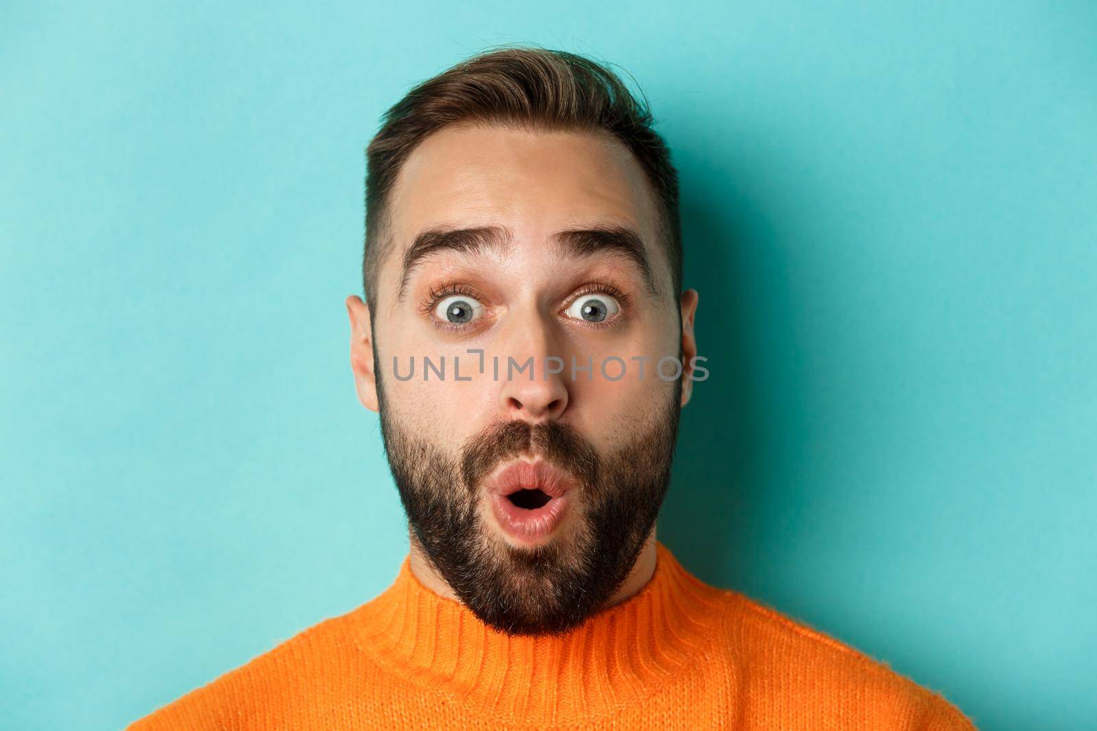 Headshot of handsome caucasian man with beard standing in orange sweater against turquoise background, saying wow and staring surprised at camera.