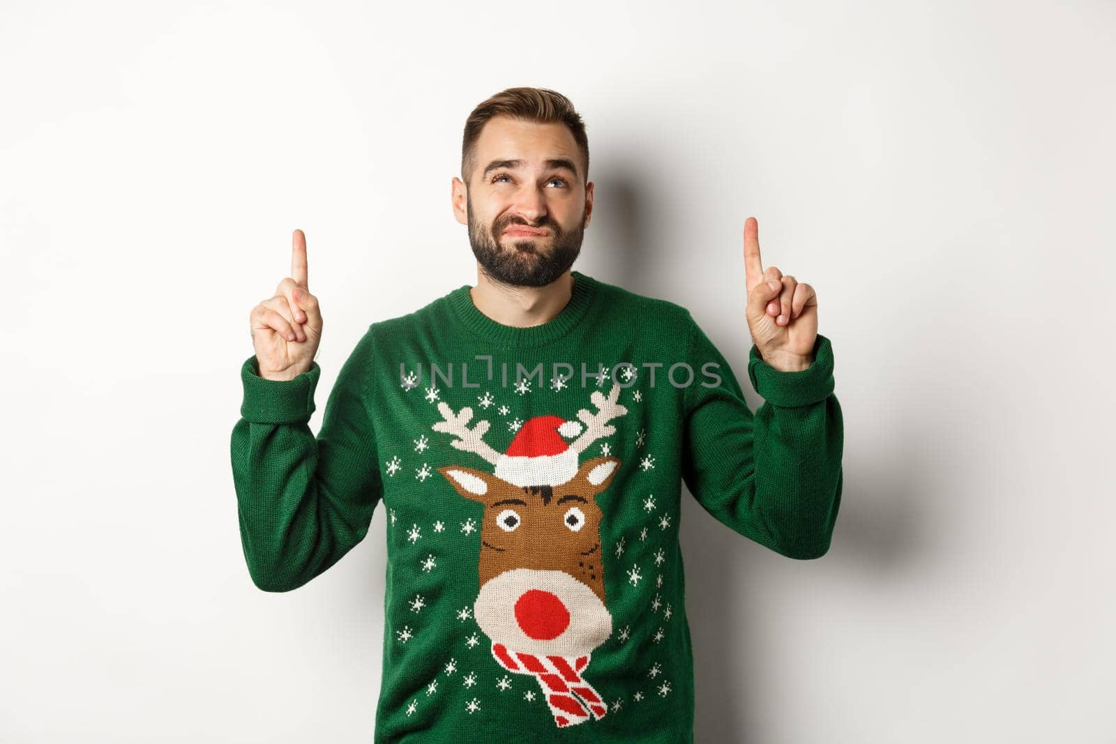 Winter holidays and christmas. Displeased guy looking doubtful at copy space, pointing fingers up and grimacing, standing over white background by Benzoix