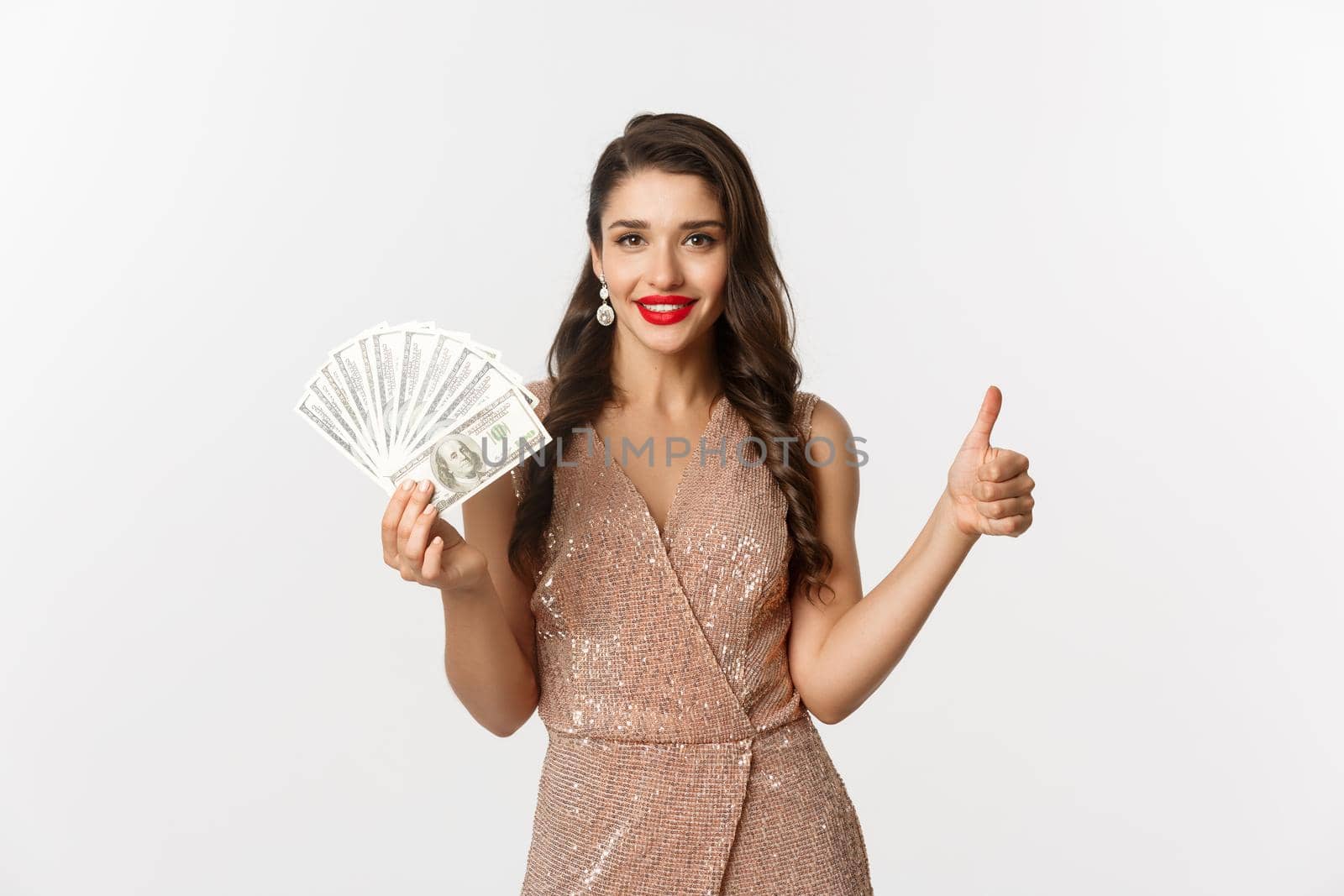 Shopping concept. Attractive, elegant woman in dress showing thumbs-up and money, recommending company, standing over white background.