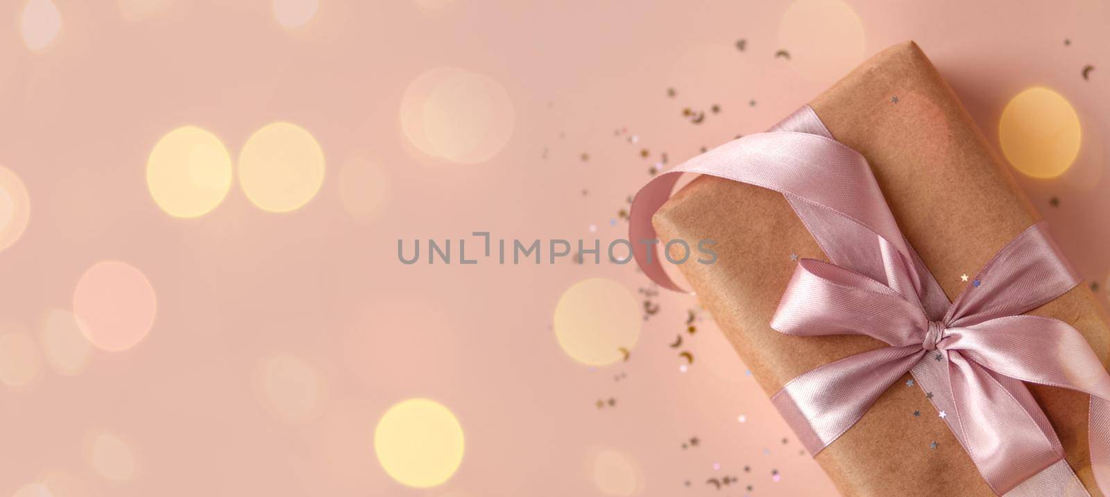 Banner of Birthday or New Year's eve present in kraft paper with pink ribbon on a soft beige background with glitter and confetti. Xmas. Flat lay. Happy holidays celebration, giving love concept