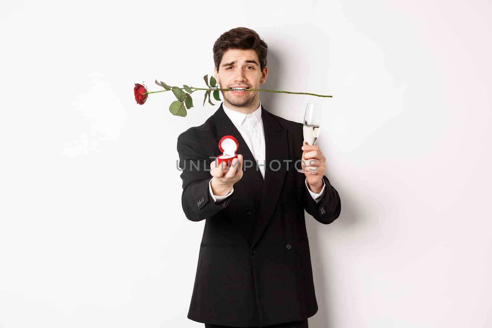 Romantic young man in suit making a proposal, holding rose in teeth and glass of champagne, showing engagement ring, asking to marry him, standing against white background by Benzoix