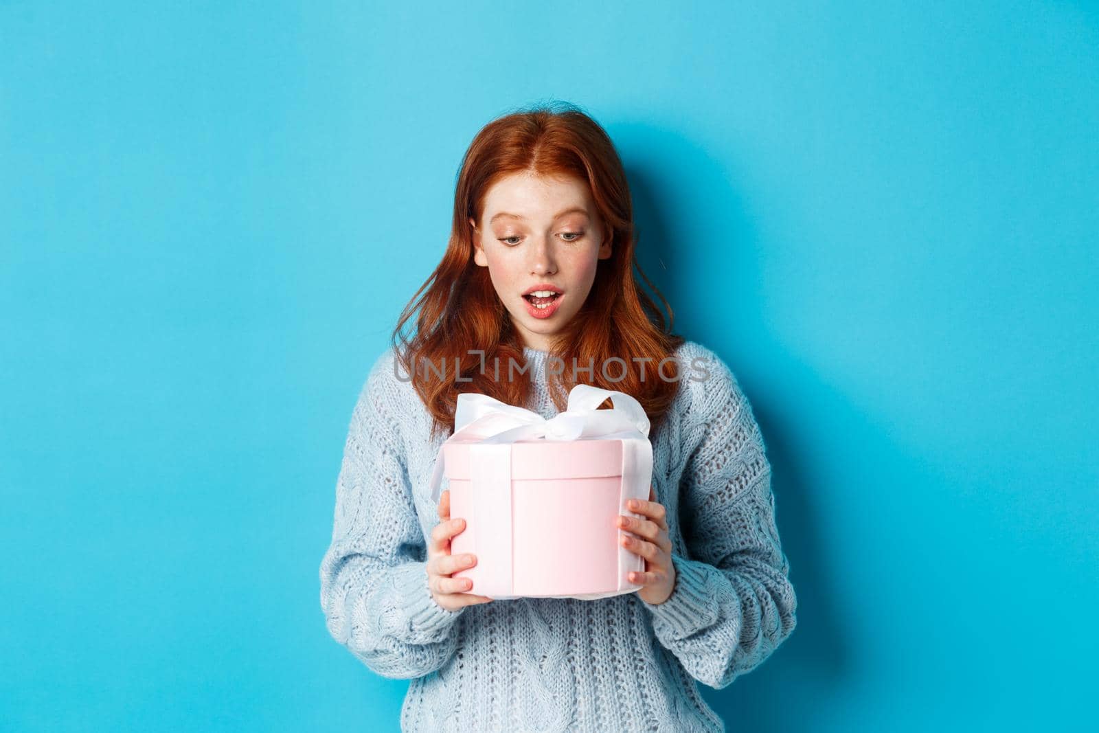 Beautiful redhead girl holding gift in pink cute box, looking at present with surprised face, standing over blue background.