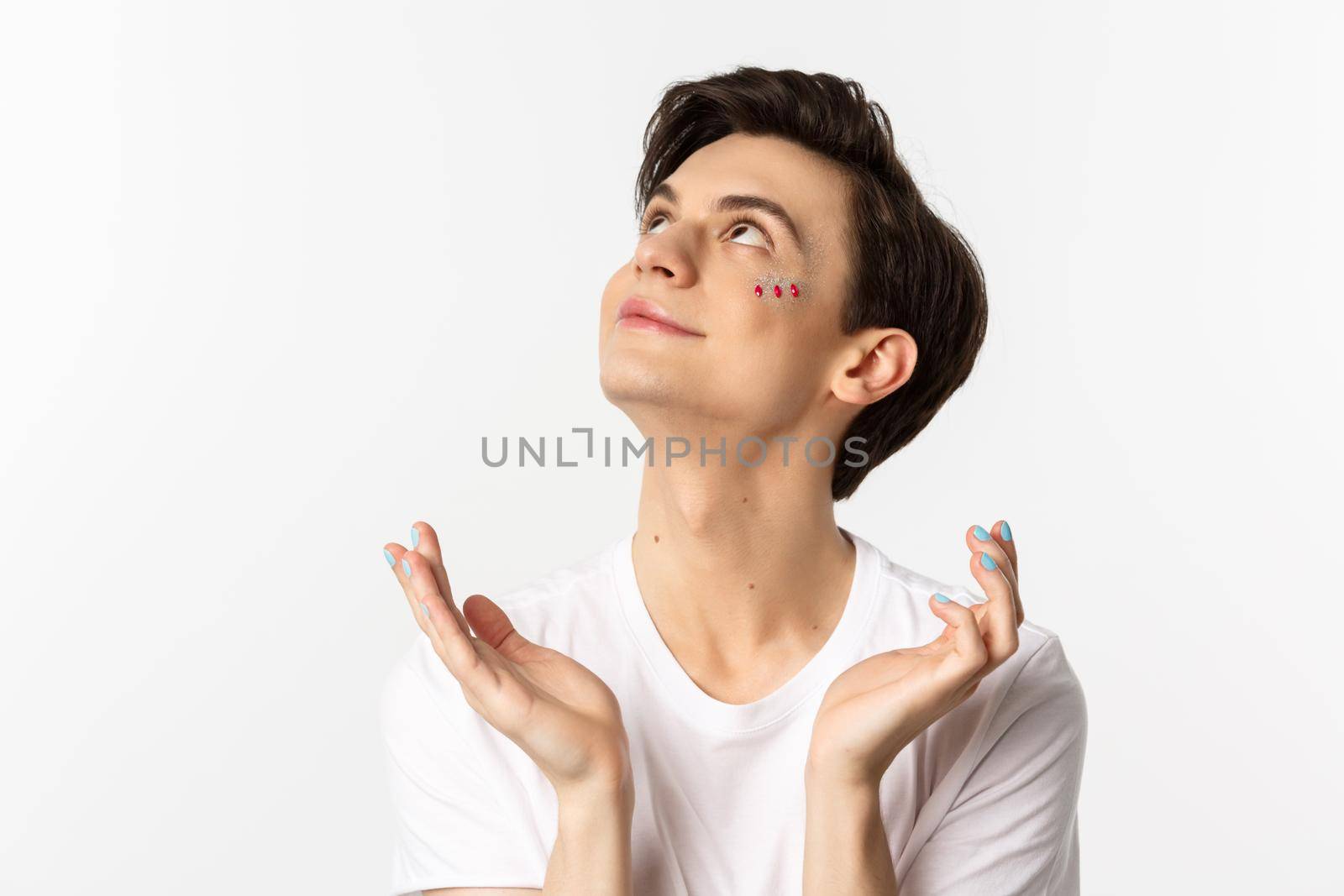 People, lgbtq and beauty concept. Close-up of handsome androgynous man with glitter on face and polished fingernails, looking dreamy up and smiling, white background.