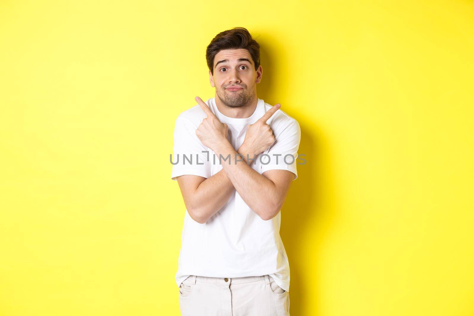 Indecisive man pointing fingers sideways, struggling to make decision, asking advice, standing over yellow background.