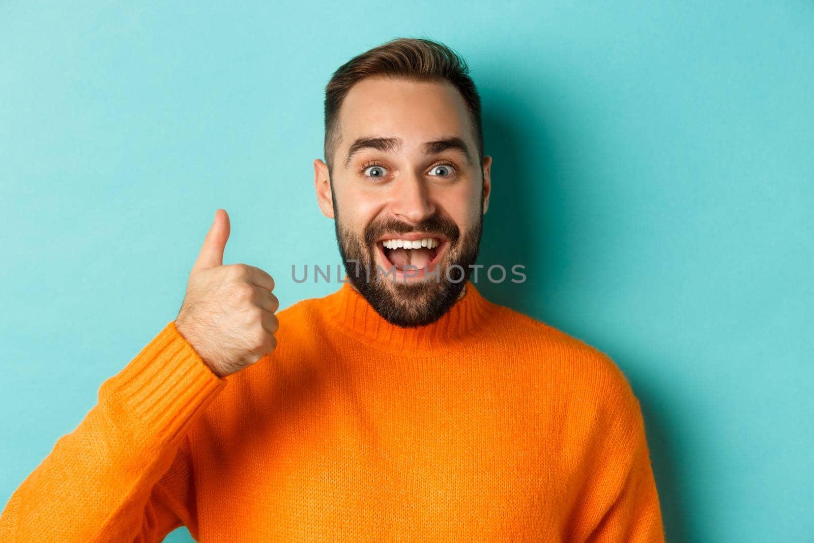 Close-up of cheerful male model showing thumb-up, smiling pleased, standing over light blue background.