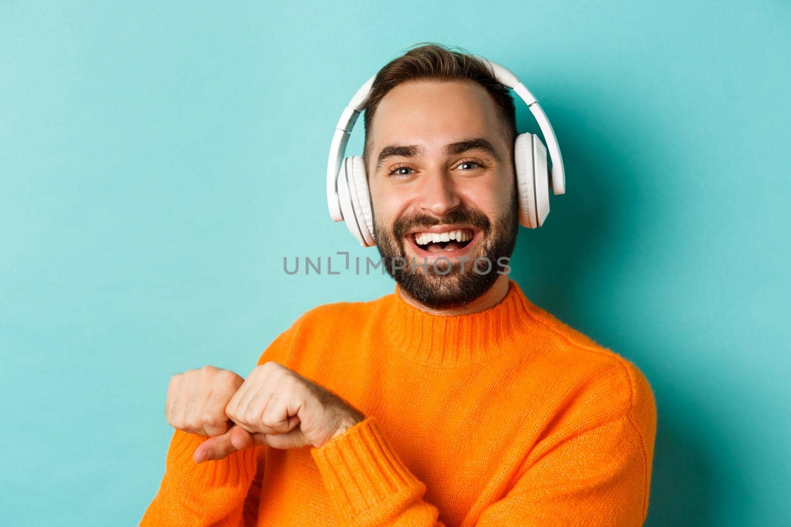 Close-up of handsome modern man listening music in headphones, standing in orange sweater over turquoise background.