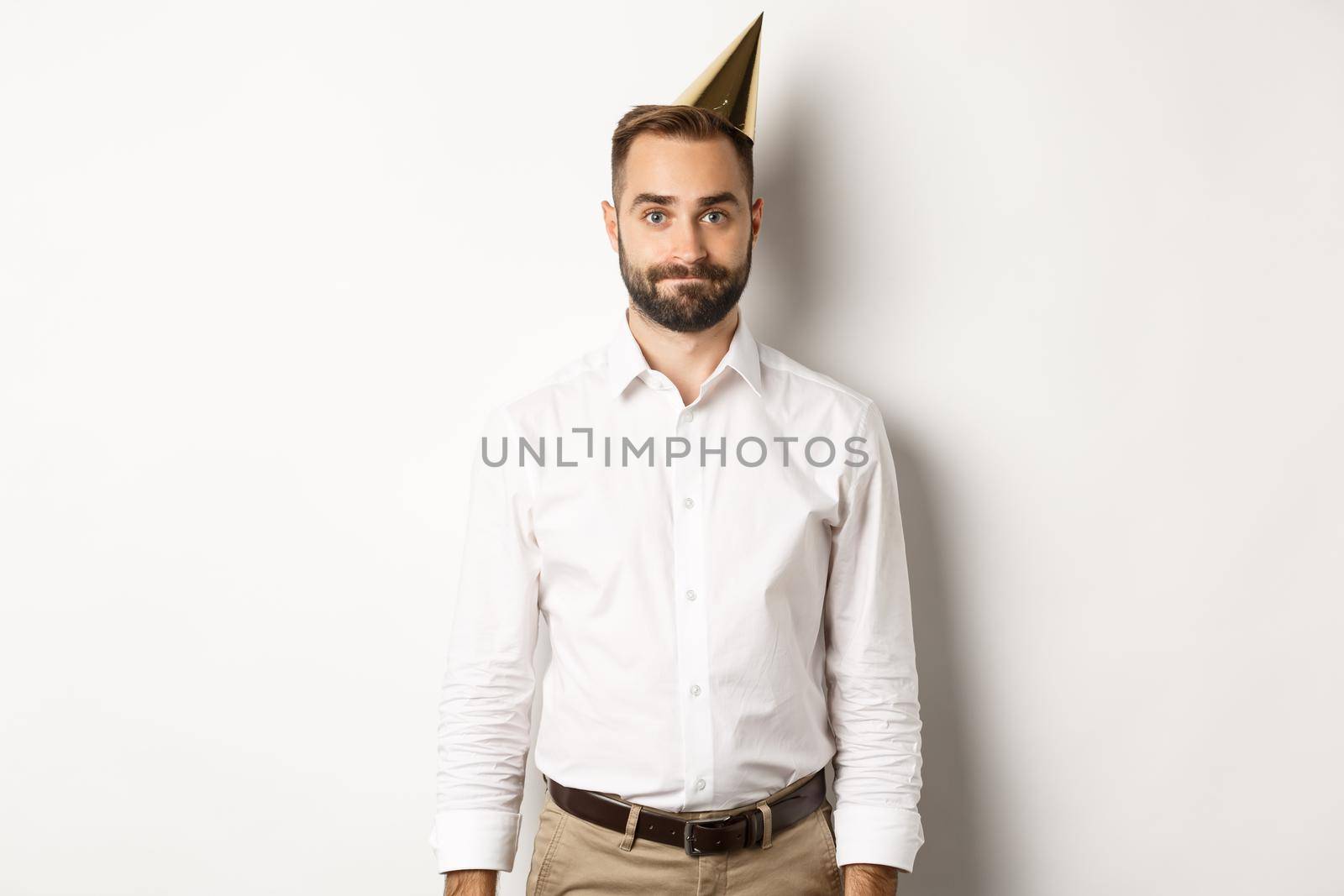 Celebration and holidays. Gloomy guy in birthday hat standing awkward against white background, feeling unamused.