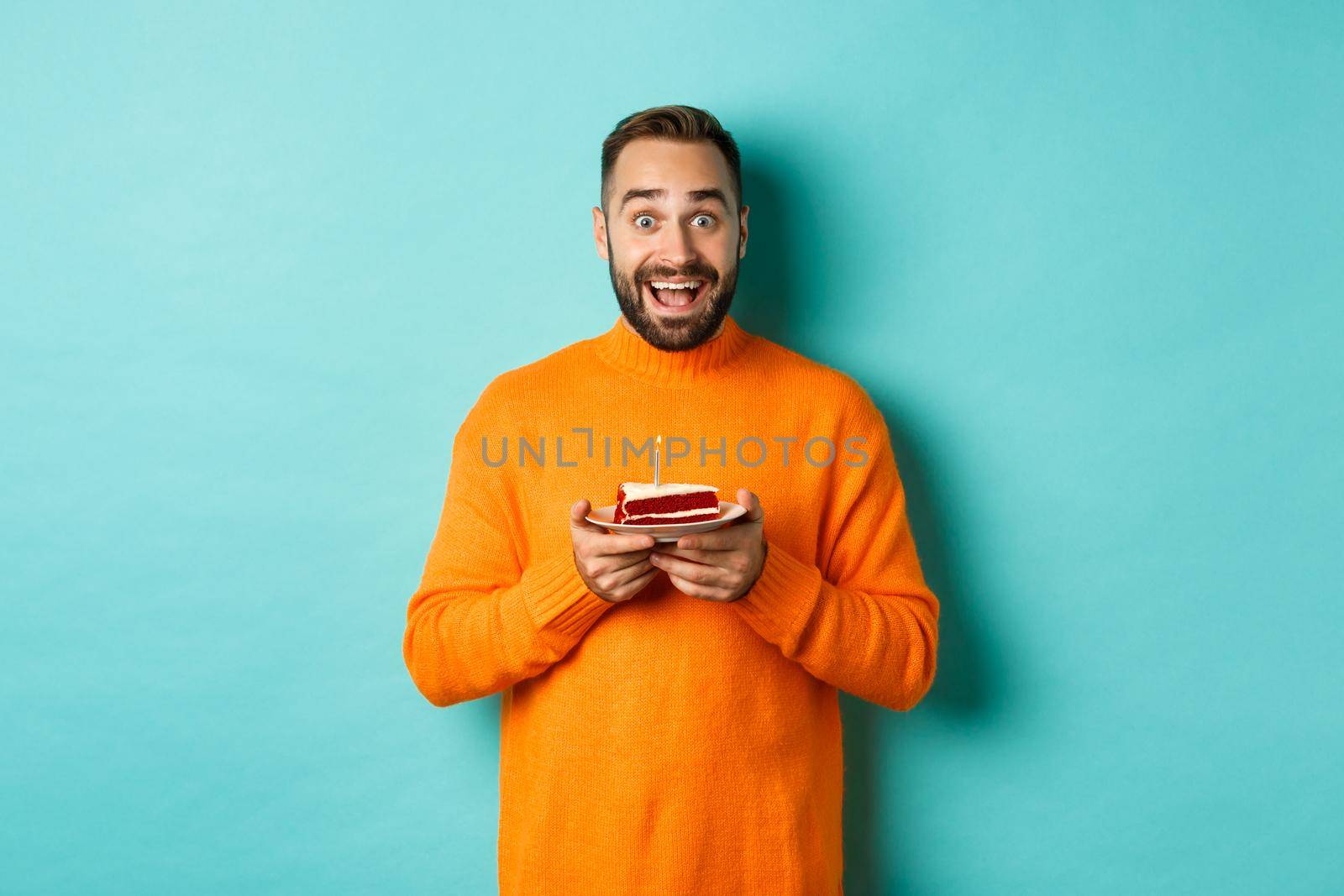 Happy adult man celebrating birthday, holding bday cake with candle and smiling, standing against turquoise background.