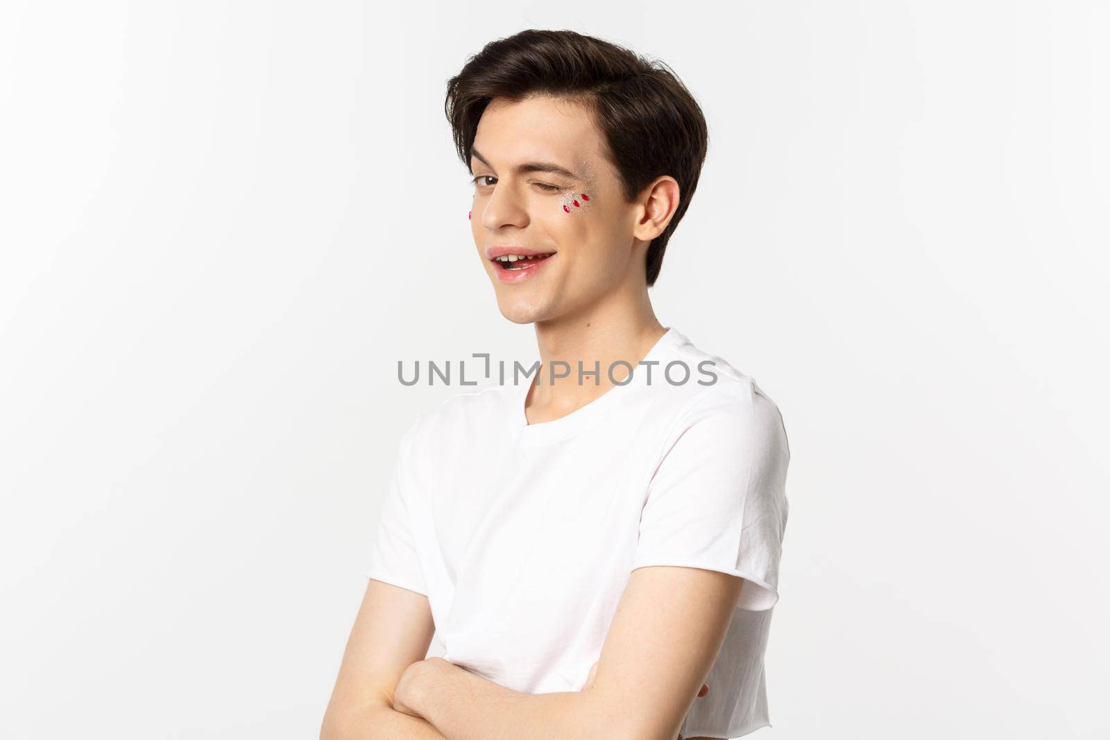 People, lgbtq and beauty concept. Beautiful cheeky gay man winking at camera and smiling, standing with glitter on face against white background.