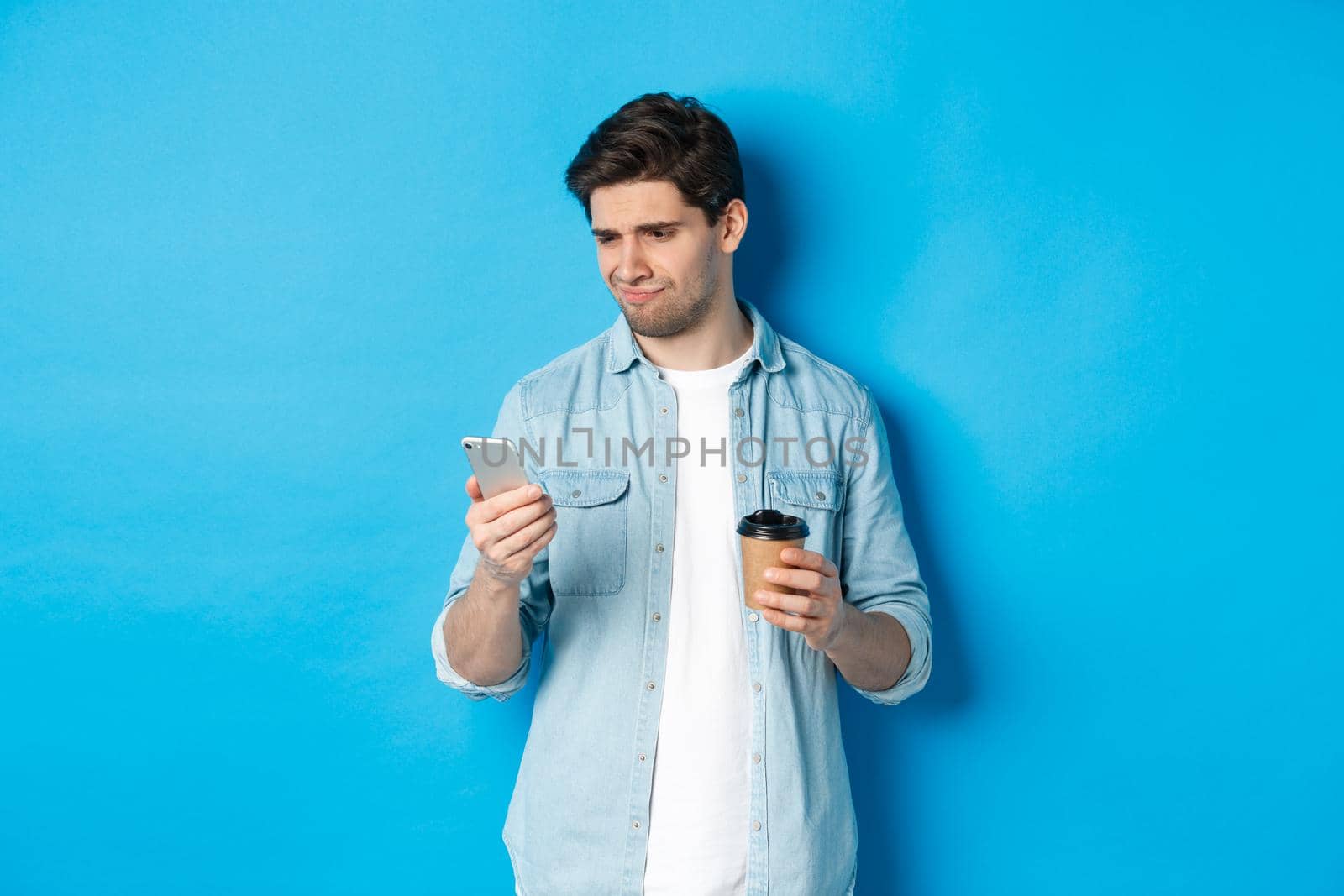 Skeptical and disappointed guy reading upsetting message on phone, holding coffee cup, standing over blue background.