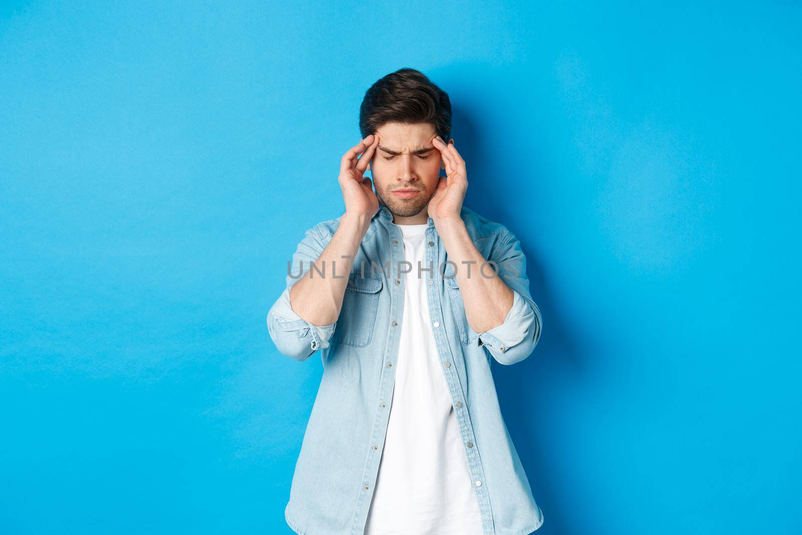 Man with hangover touching head and grimacing, having headache, standing against blue background.