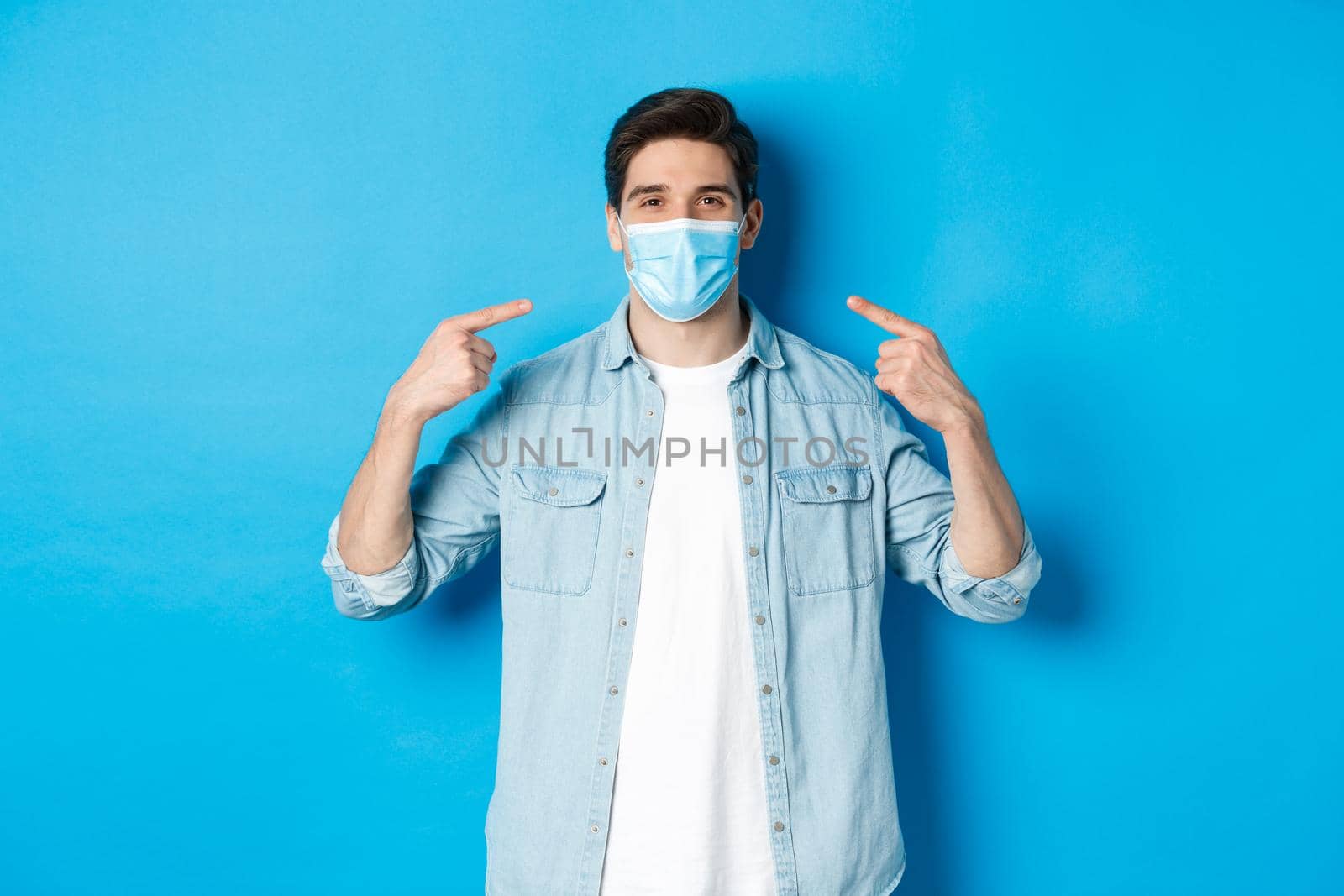 Concept of coronavirus, quarantine and social distancing. Handsome man pointing at medical mask and smiling, protection from virus spread during pandemic, blue background by Benzoix