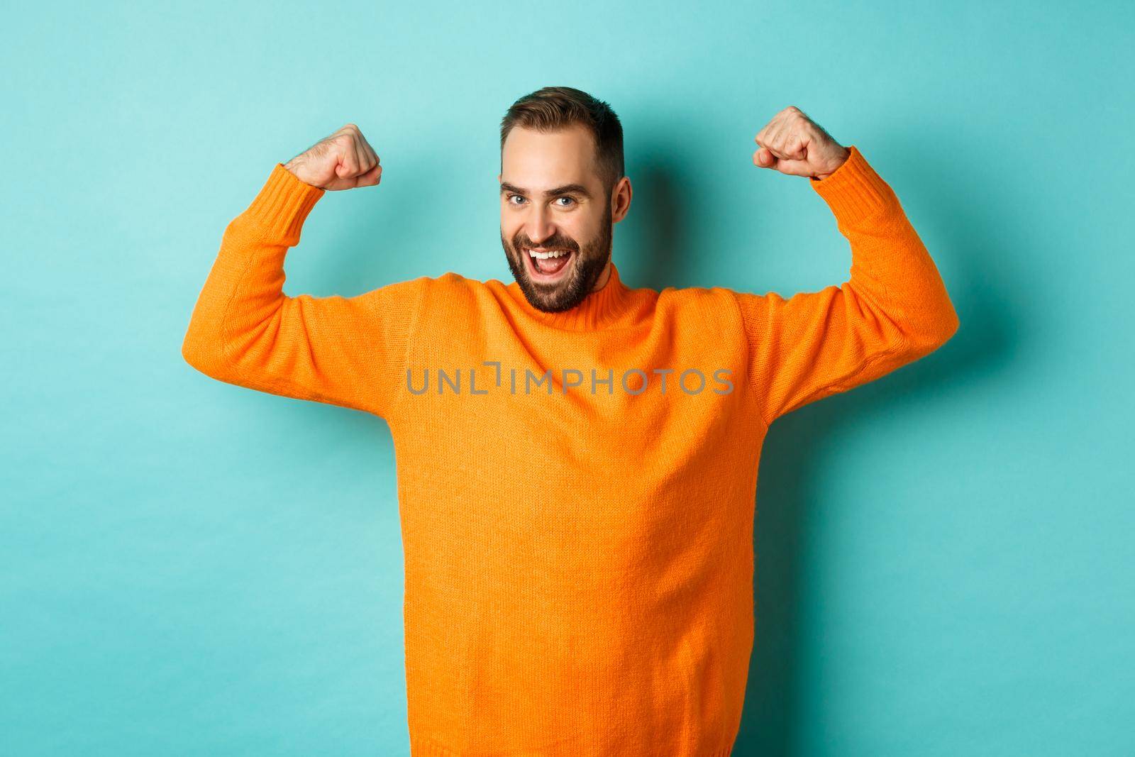 Young and strong man looking confident, flex biceps and show-off muscle strengths, smiling self-assured, standing over light blue background.