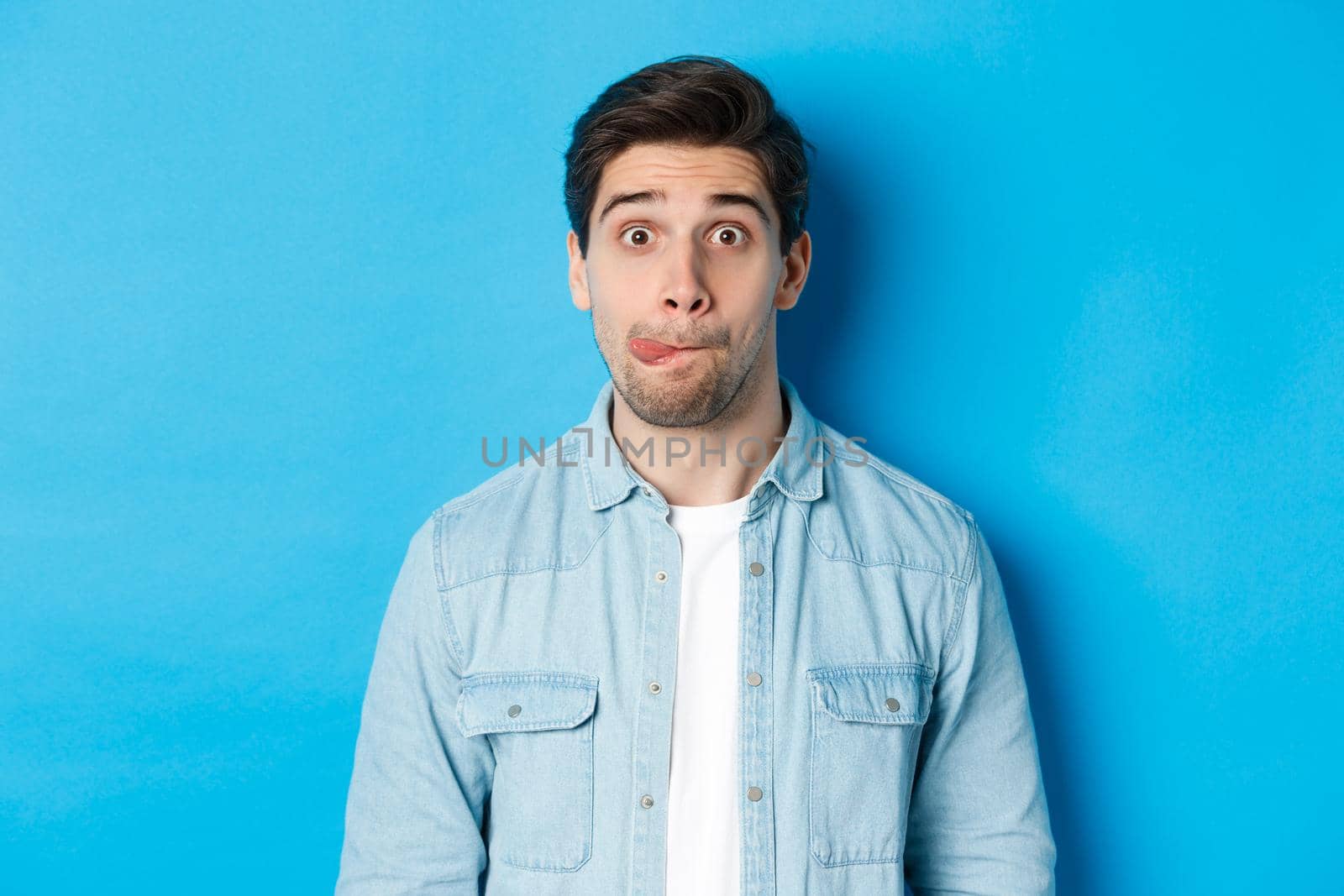 Close-up of young man making funny expressions, showing tongue and looking at camera, standing over blue background.